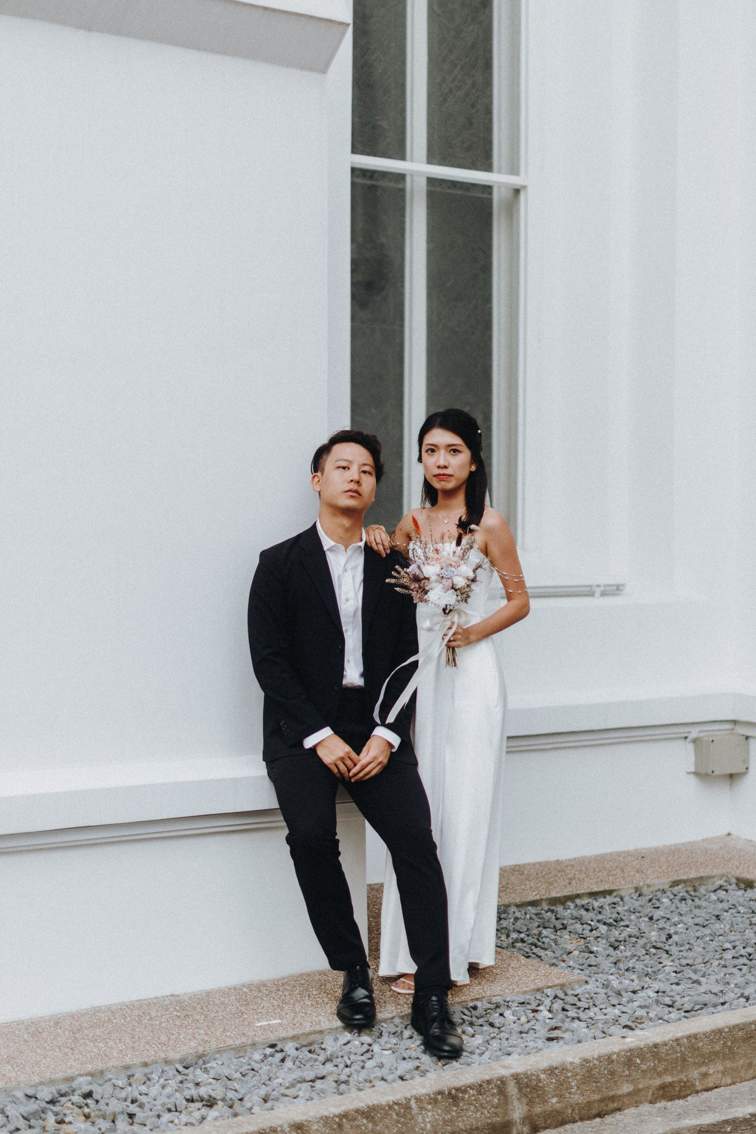 A couple in wedding attire poses against a white wall with large windows.