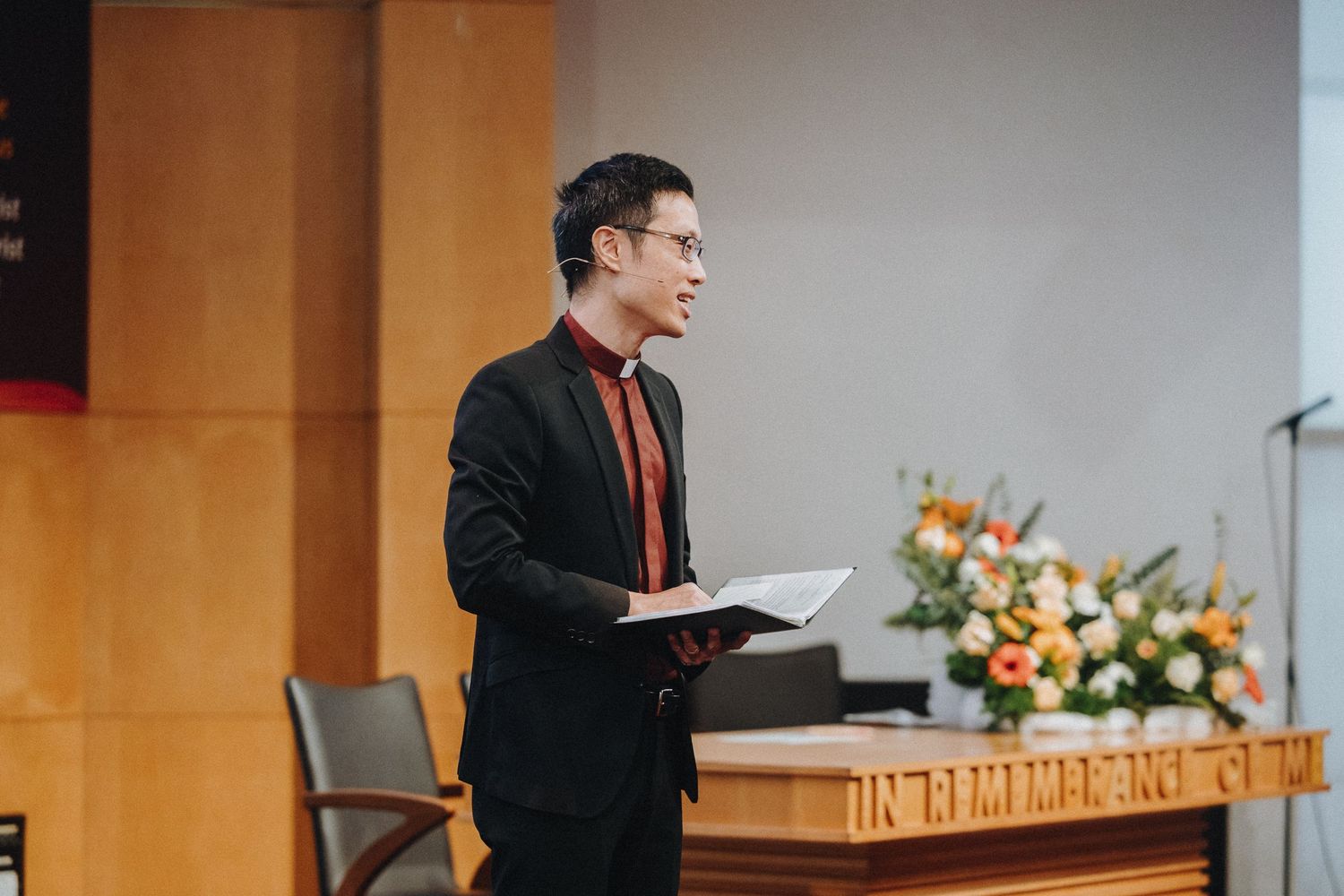 A speaker in formal attire delivers a presentation at a podium with floral decorations.