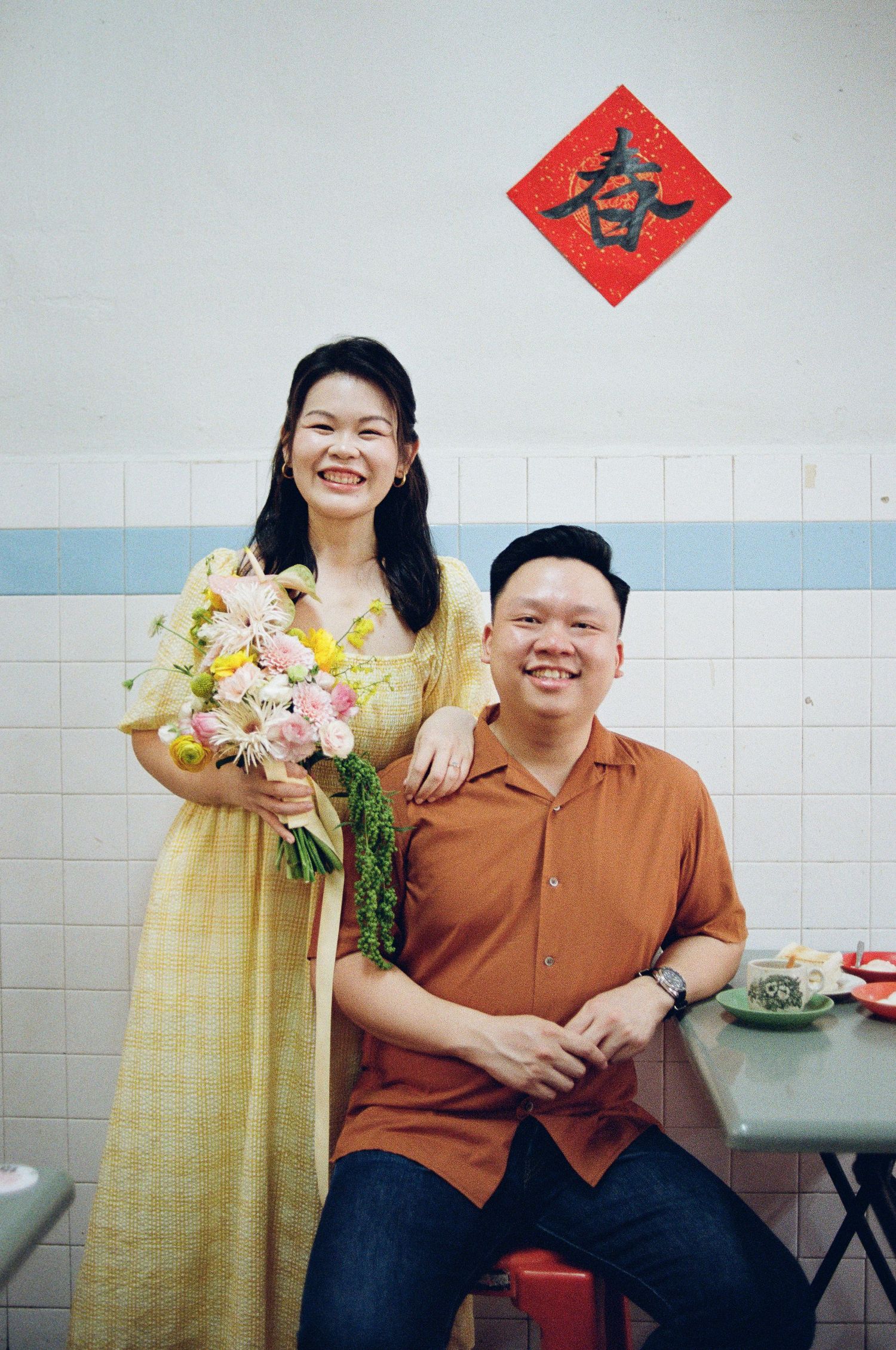 A couple poses together in a bright interior with a red Chinese decoration on the wall and flowers in yellow and pink tones.