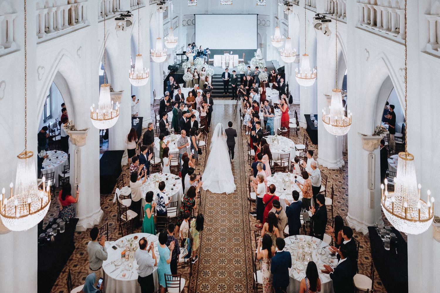 Aerial view of an elegant wedding reception in a grand white hall with chandeliers, guests at round tables, and bride and groom walking.