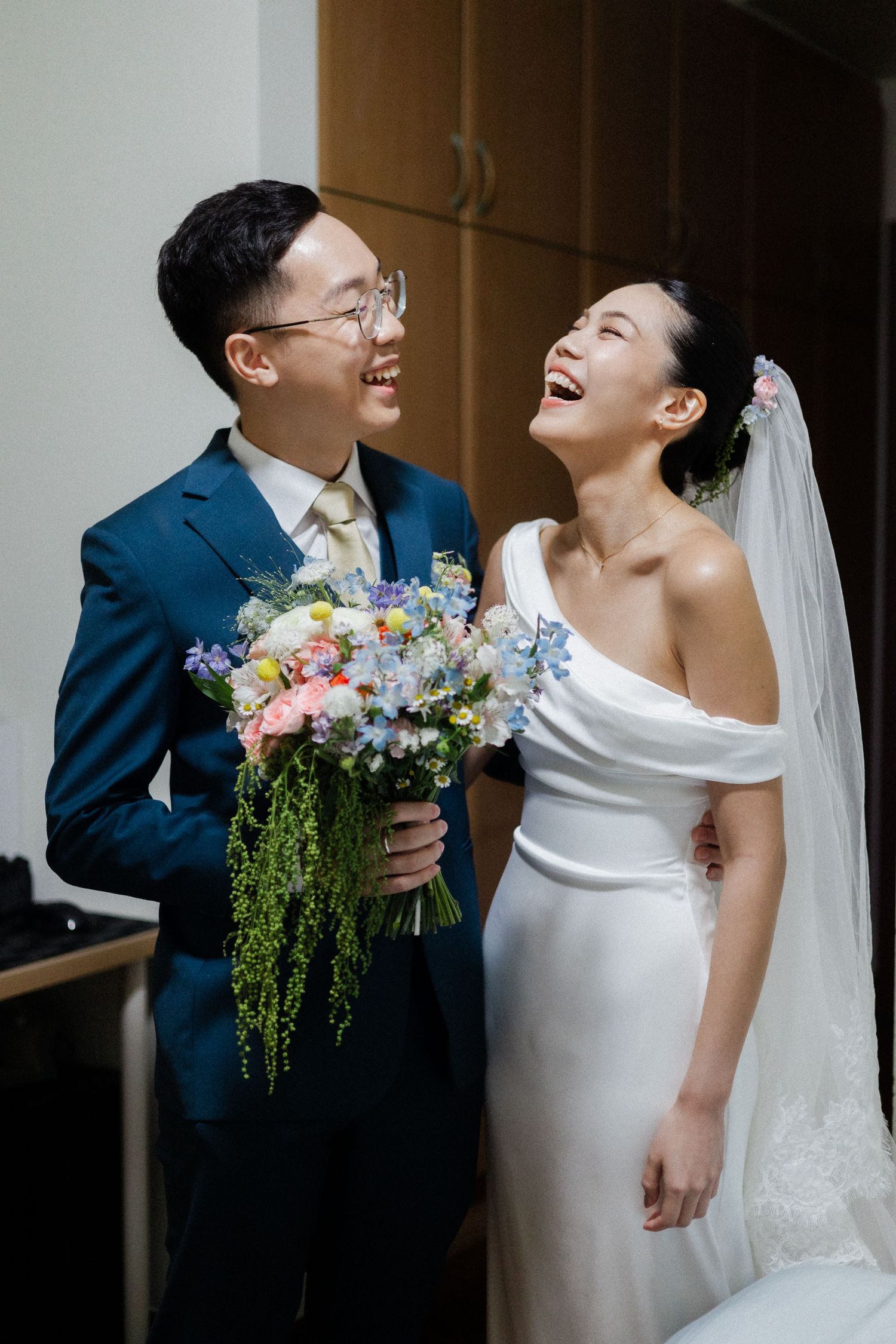 Joyful Asian bride and groom laughing together, bride in white off-shoulder gown with veil, groom in navy suit holding colorful bouquet.