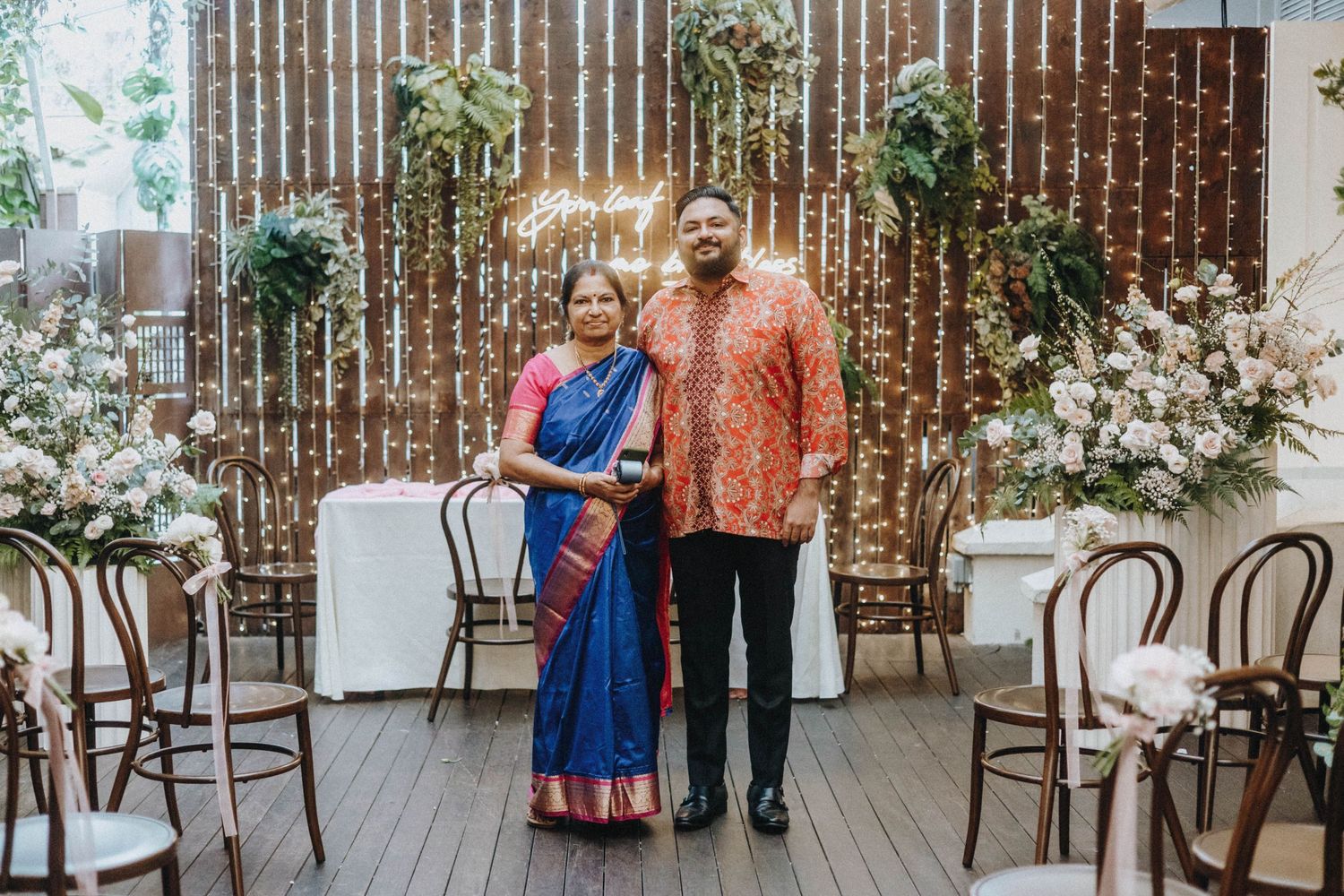 Couple in traditional Indian attire posing at an elegant wedding venue with floral decor and fairy lights.