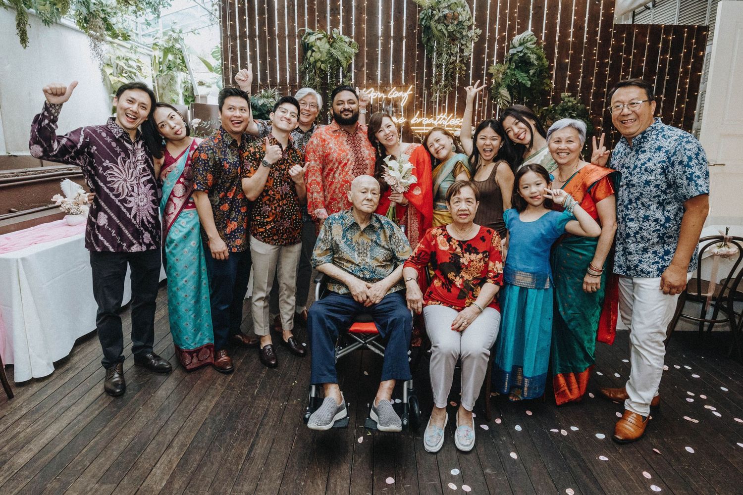 A large multicultural wedding group photo taken outdoors with an elderly guest seated in a wheelchair.