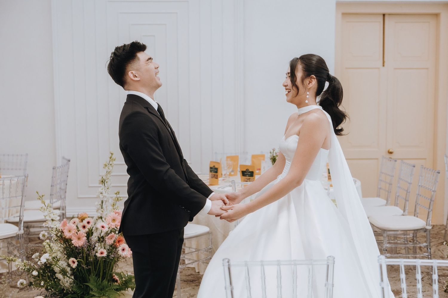 Bride and groom hold hands and smile at each other during their wedding ceremony in a bright white venue.