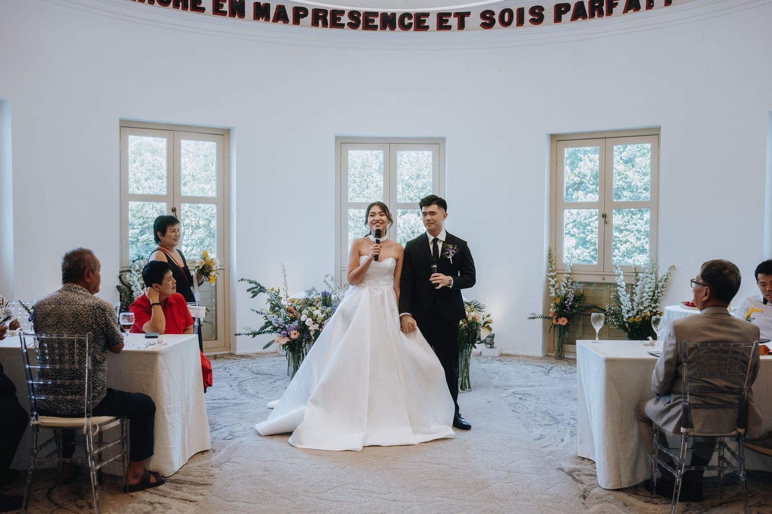 Bride speaks into microphone during intimate wedding ceremony while groom stands beside her.