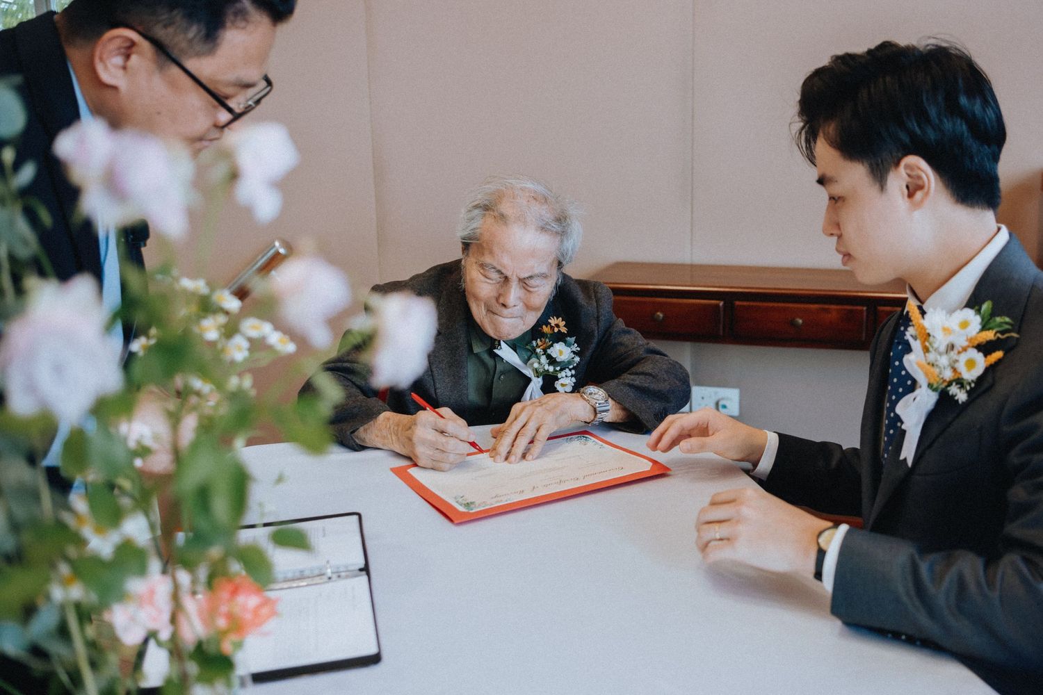 Elderly officiant signs wedding documents at ceremony signing table with groom.