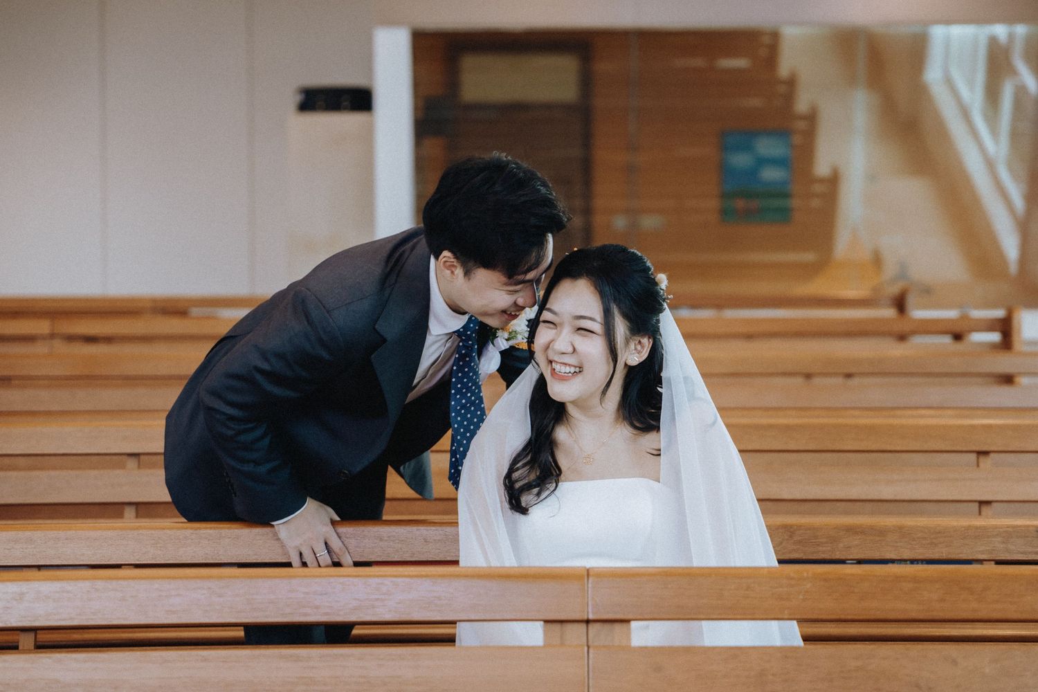 A smiling bride in a white gown and veil sits in church pews as a groom in a dark suit leans in close.