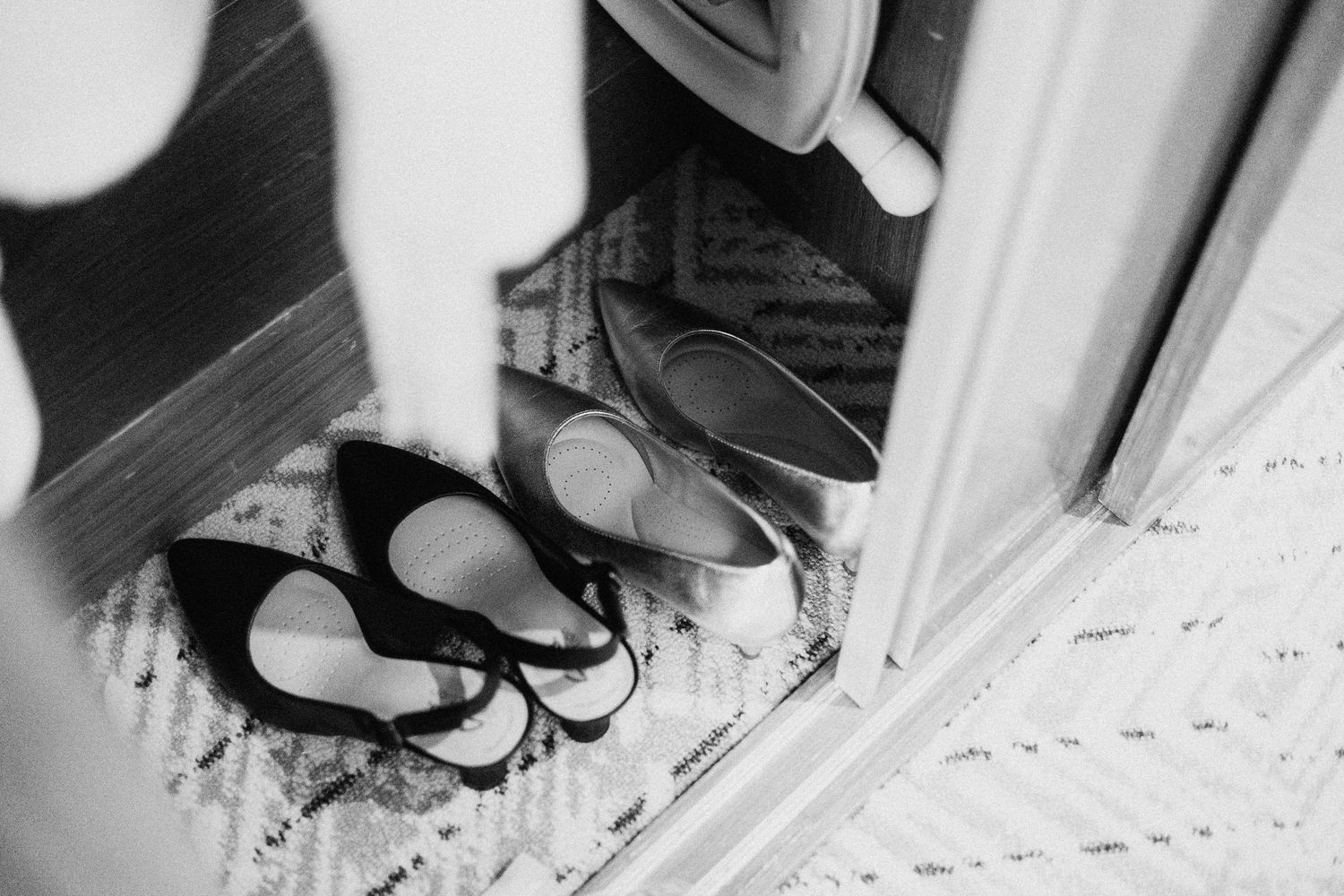 Black and white photo of elegant women's heels arranged neatly by a doorway on a patterned rug.