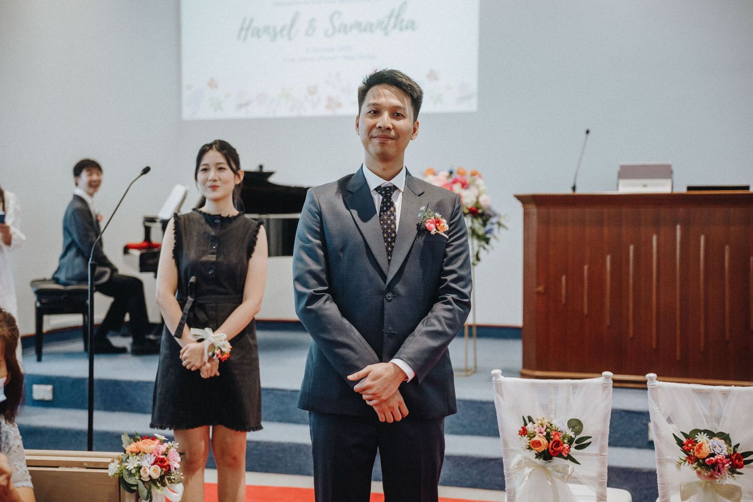 A groom in grey suit stands at church altar with wedding decorations and guests in background.