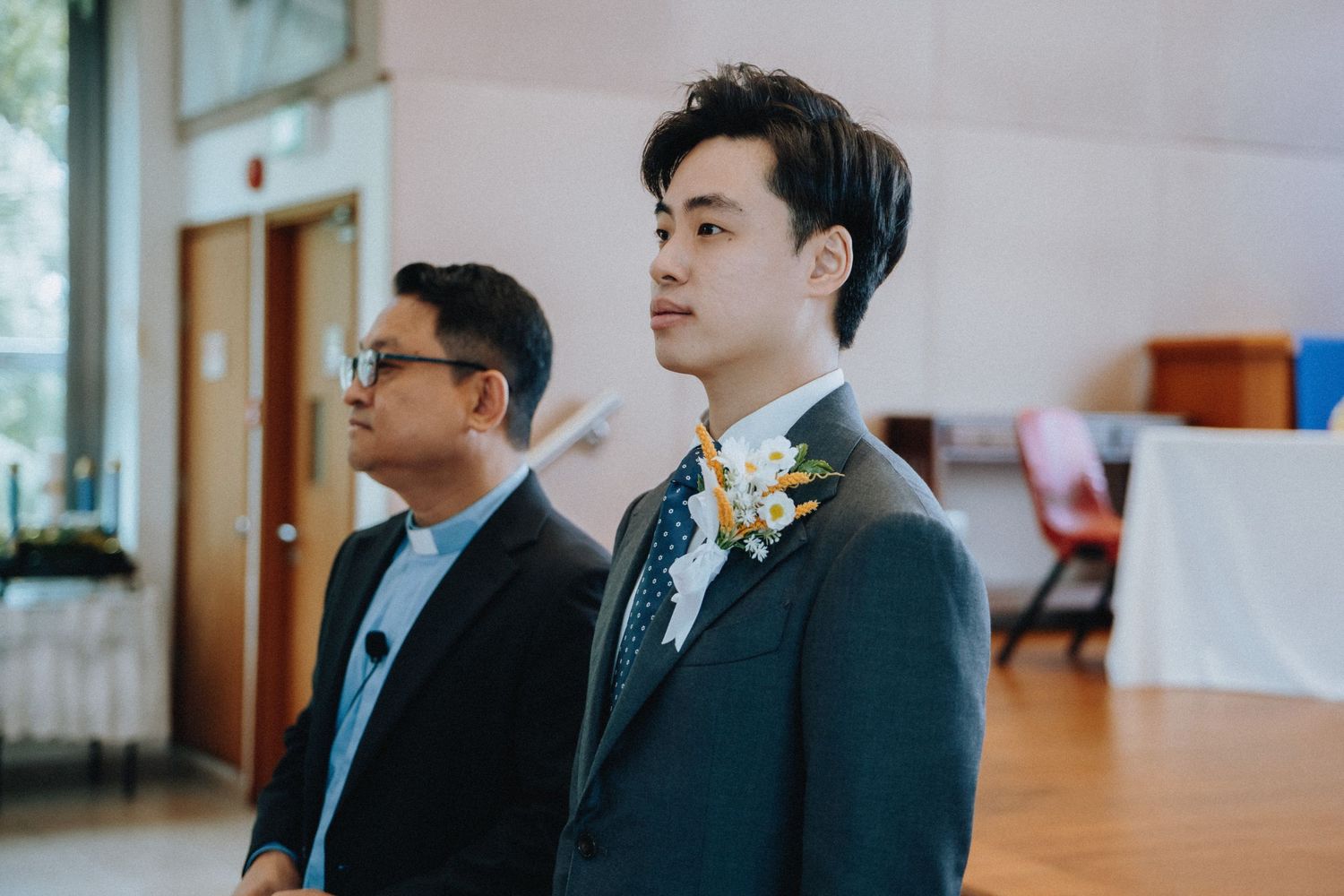 Groom in dark suit with boutonniere stands solemnly at wedding ceremony beside older man in glasses.