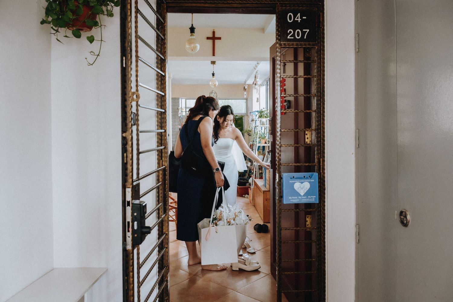 Bride and bridesmaid preparing at apartment doorway with wedding gifts and bags on the floor.