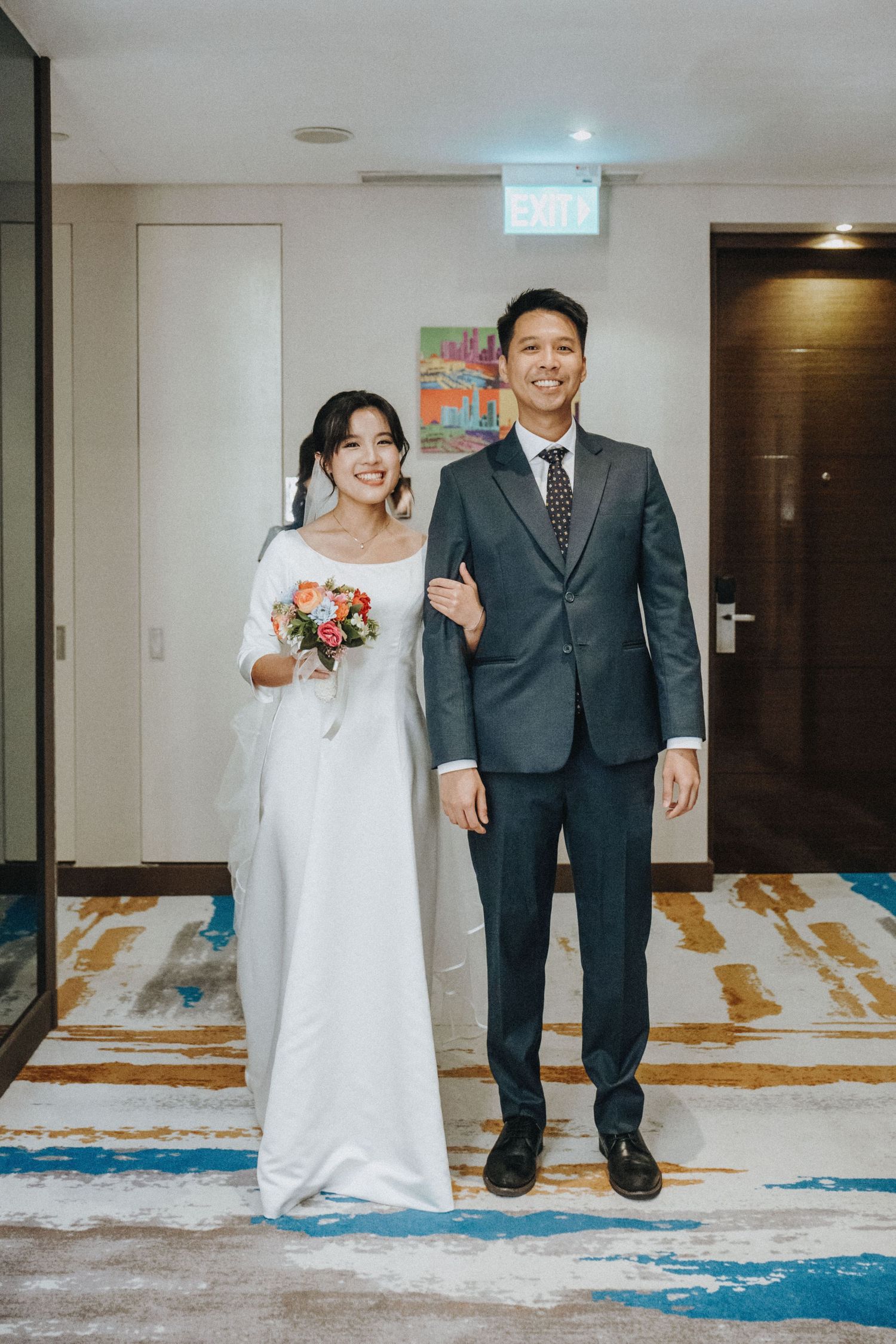 A smiling bride in a white gown holds a bouquet beside a groom in a gray suit in a hotel hallway.