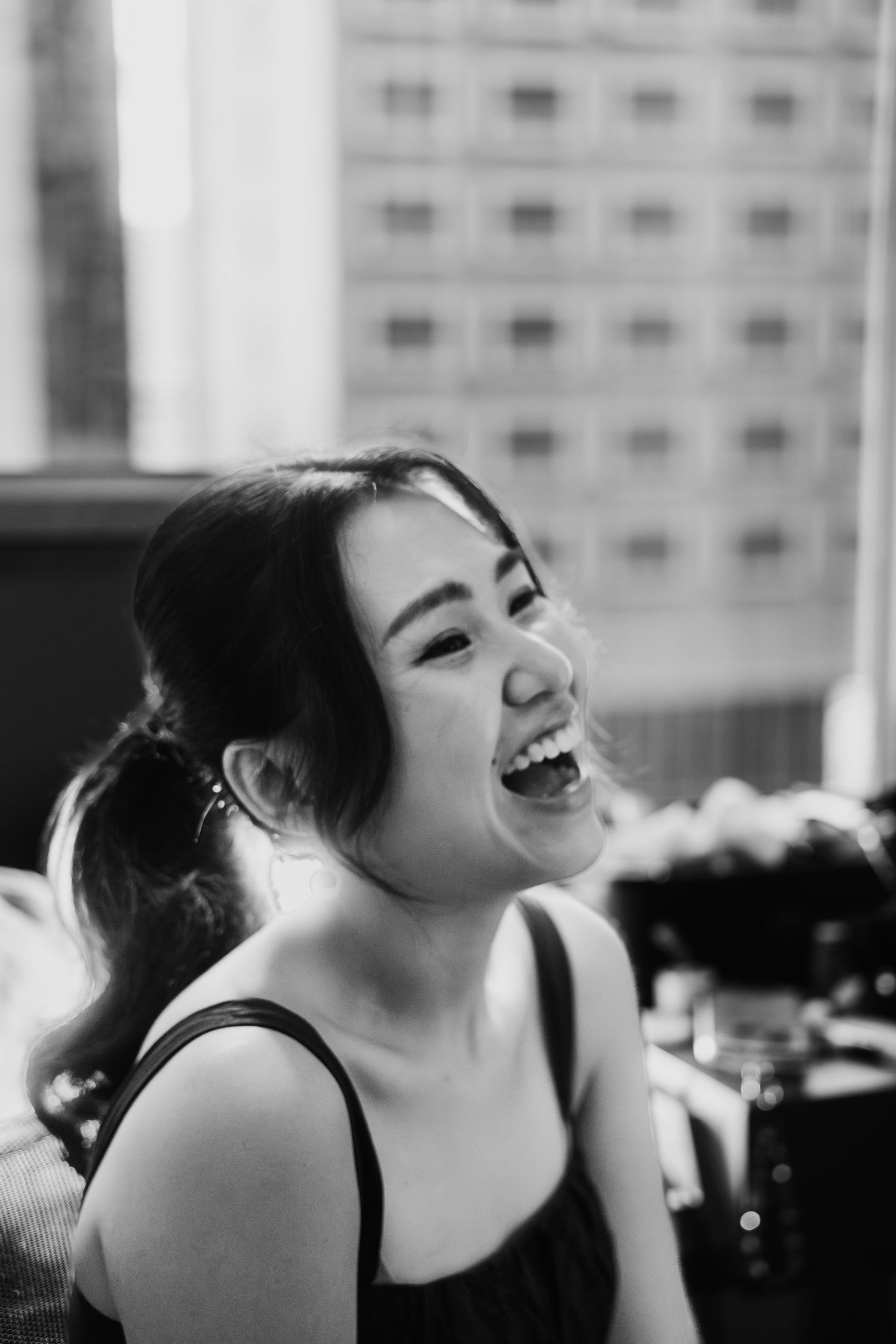 Black and white portrait of a young Asian woman laughing joyfully outdoors with a city building in the background.
