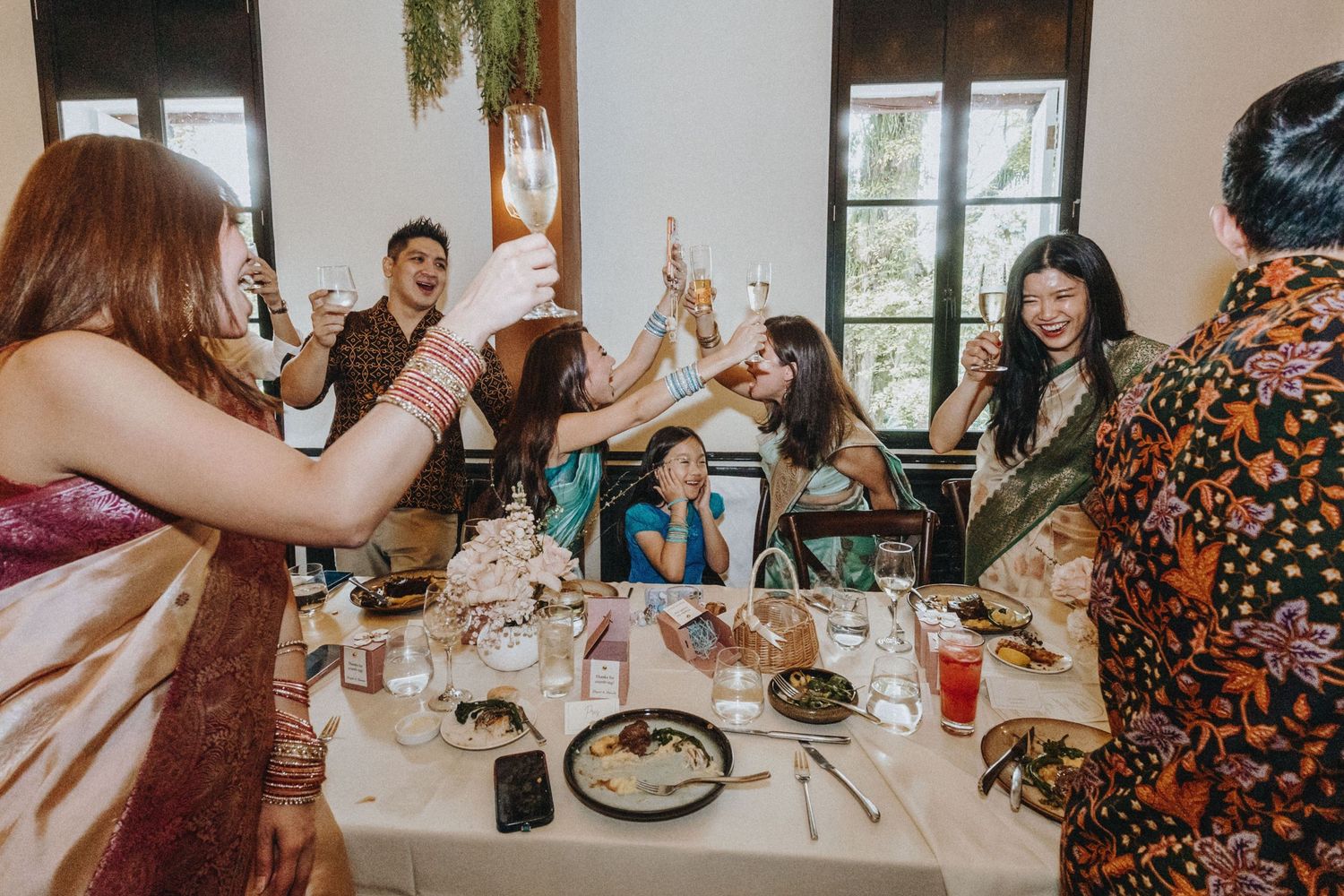 Wedding guests raise glasses in a toast at a festive indoor reception dinner table.