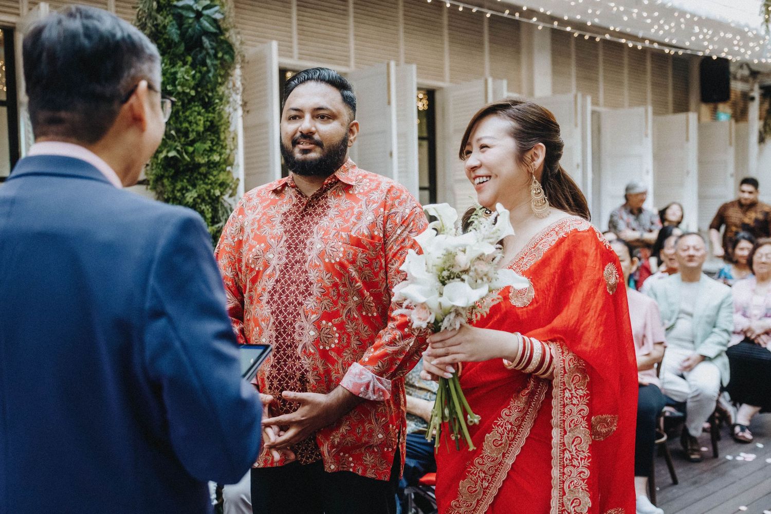 Bride in red saree holds white flowers during multicultural wedding ceremony outdoors.