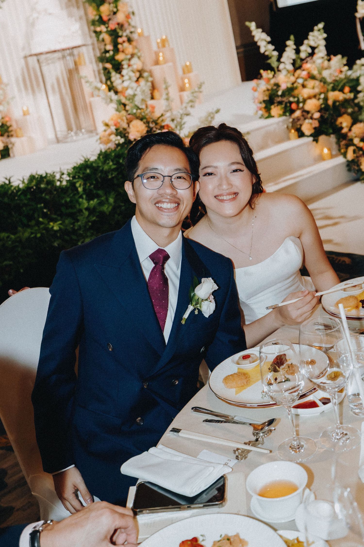 A couple dining at an elegant wedding reception with floral arrangements and place settings at a white-clothed table.