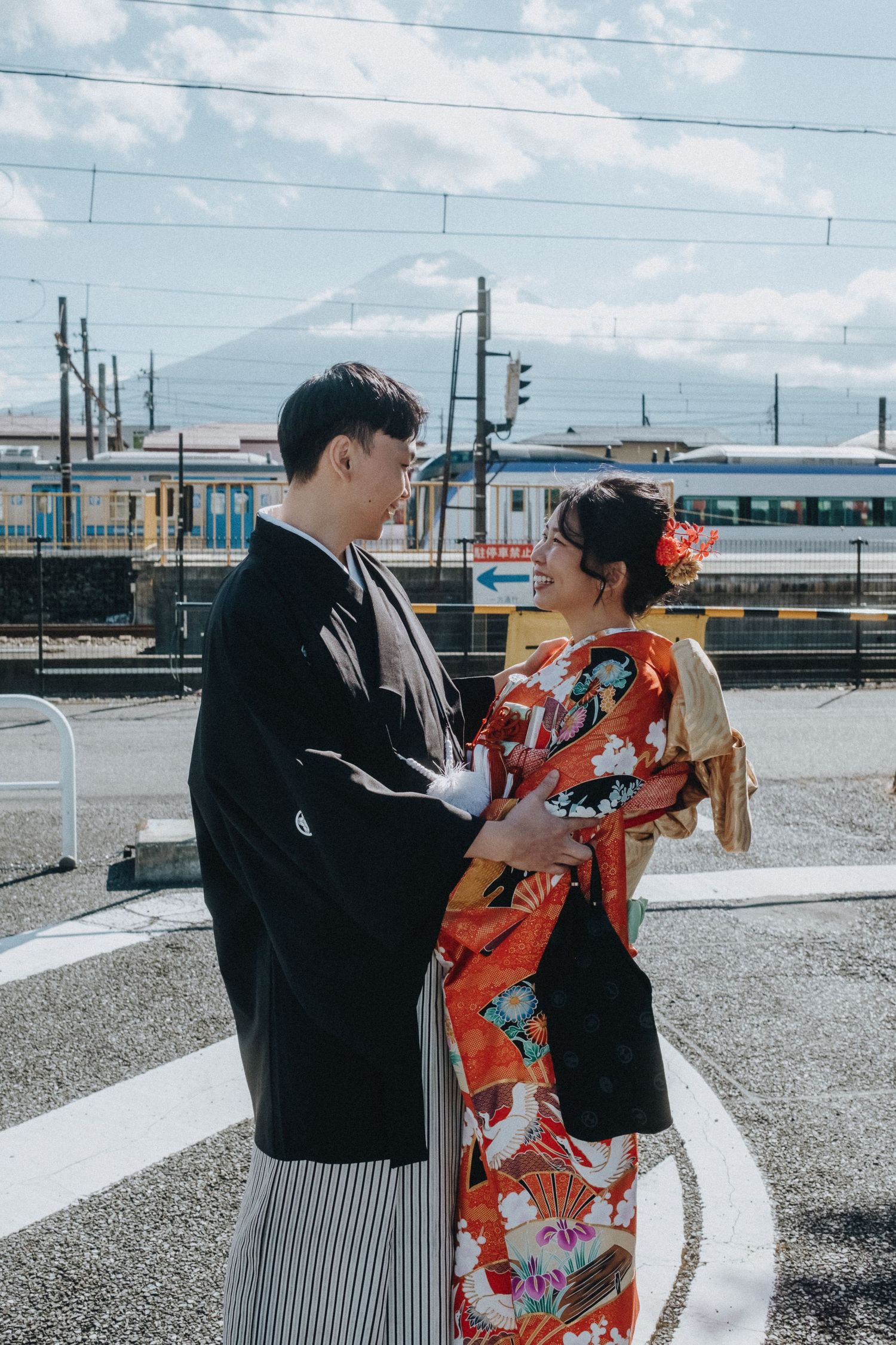 A couple in traditional Japanese attire poses against an industrial backdrop with power lines and mountains.