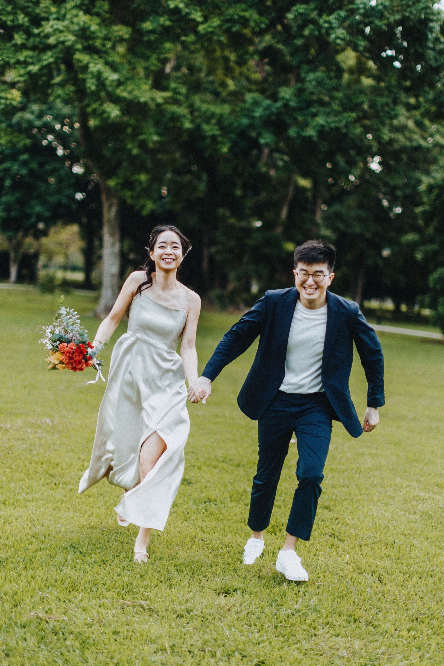 A couple runs playfully across a green lawn, with the bride in white and groom in navy during their wedding photoshoot.
