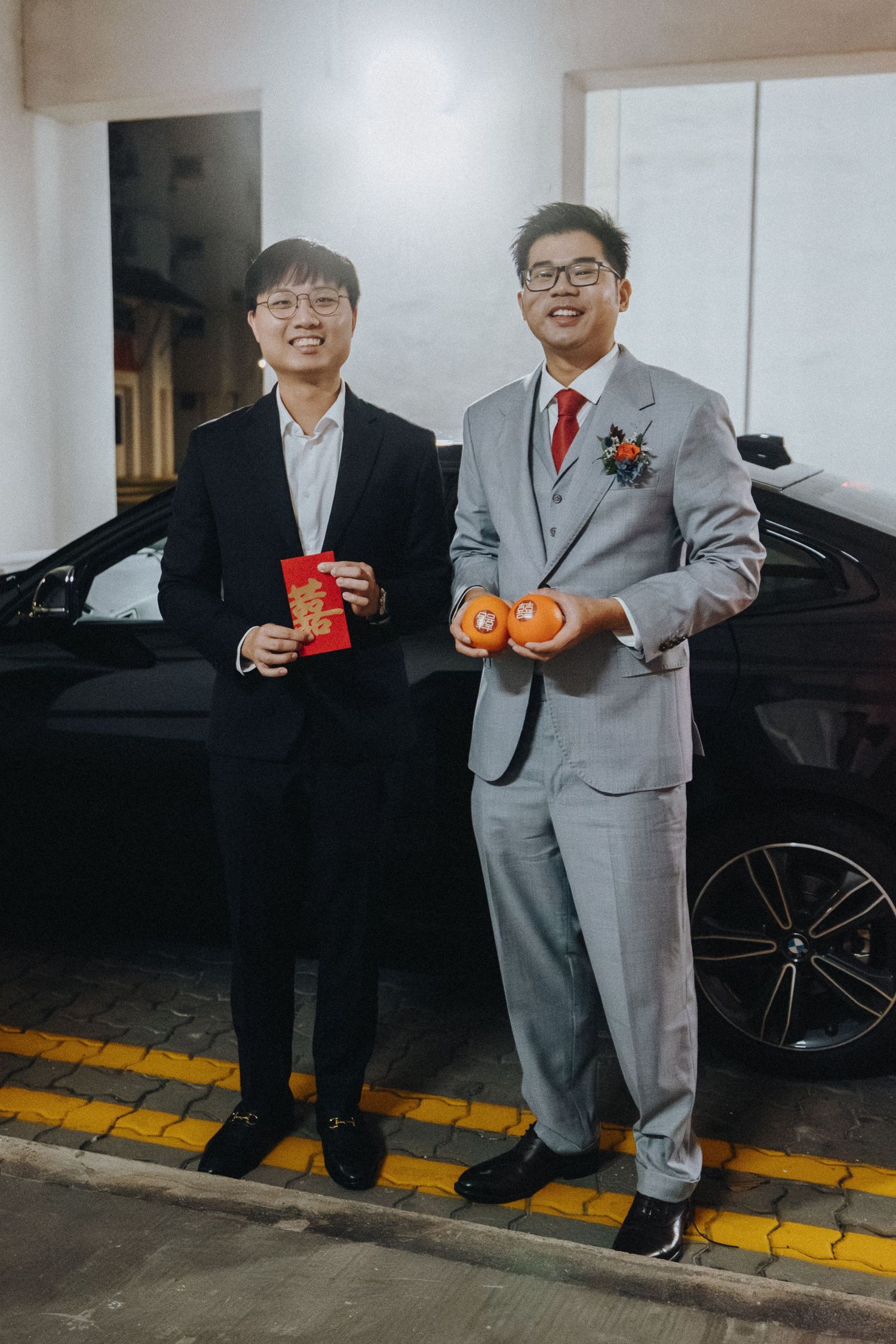 Wedding party members in gray suits hold red envelopes and oranges in a parking garage.