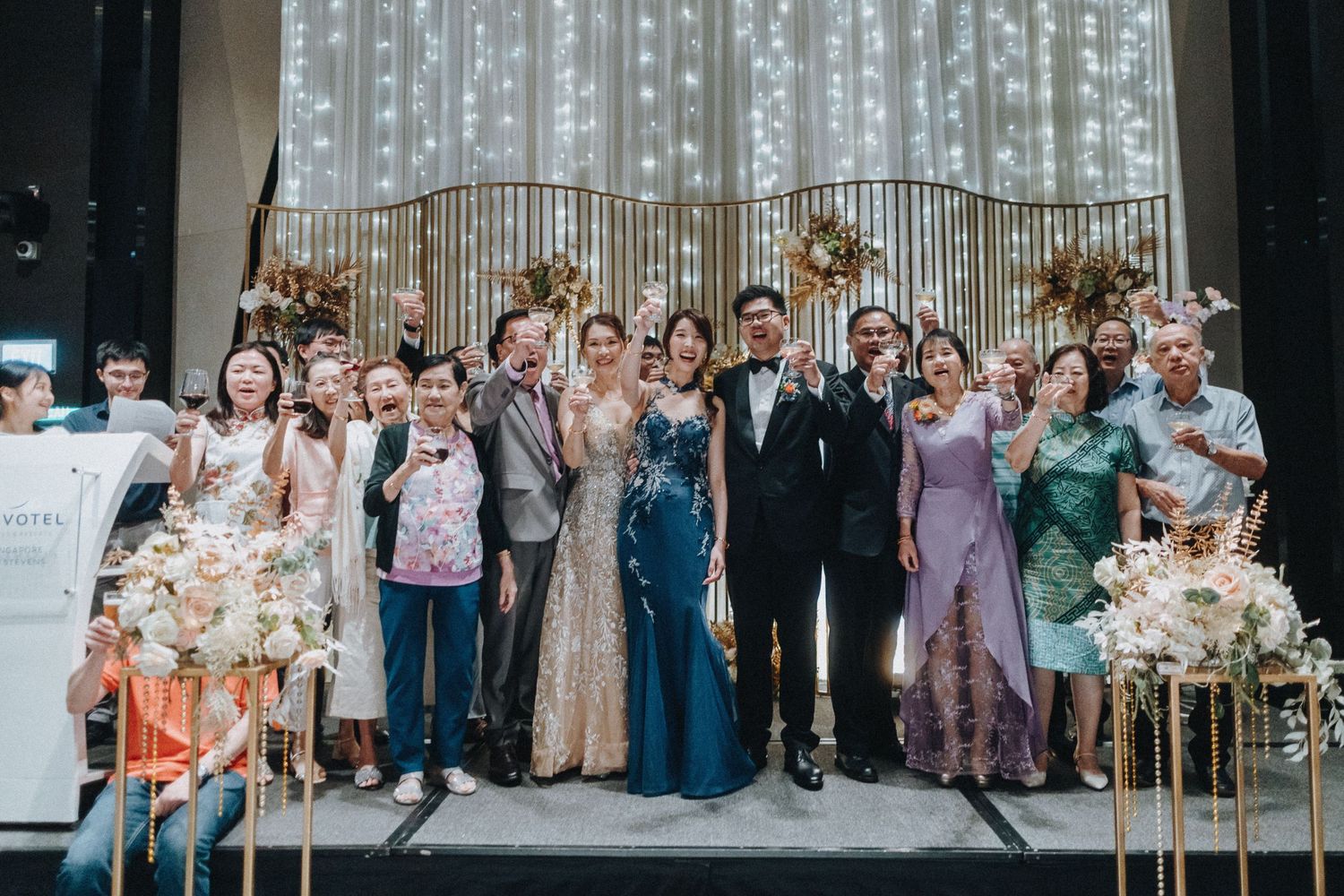 Wedding party group poses with bouquets outdoors in formal attire near grand windows.