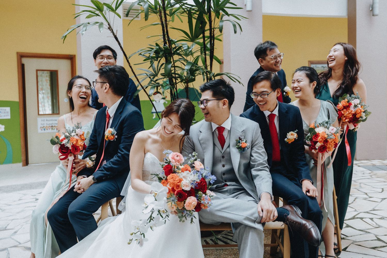 Wedding party sits on outdoor bench sharing joyful laughter with colorful bouquets of red flowers.