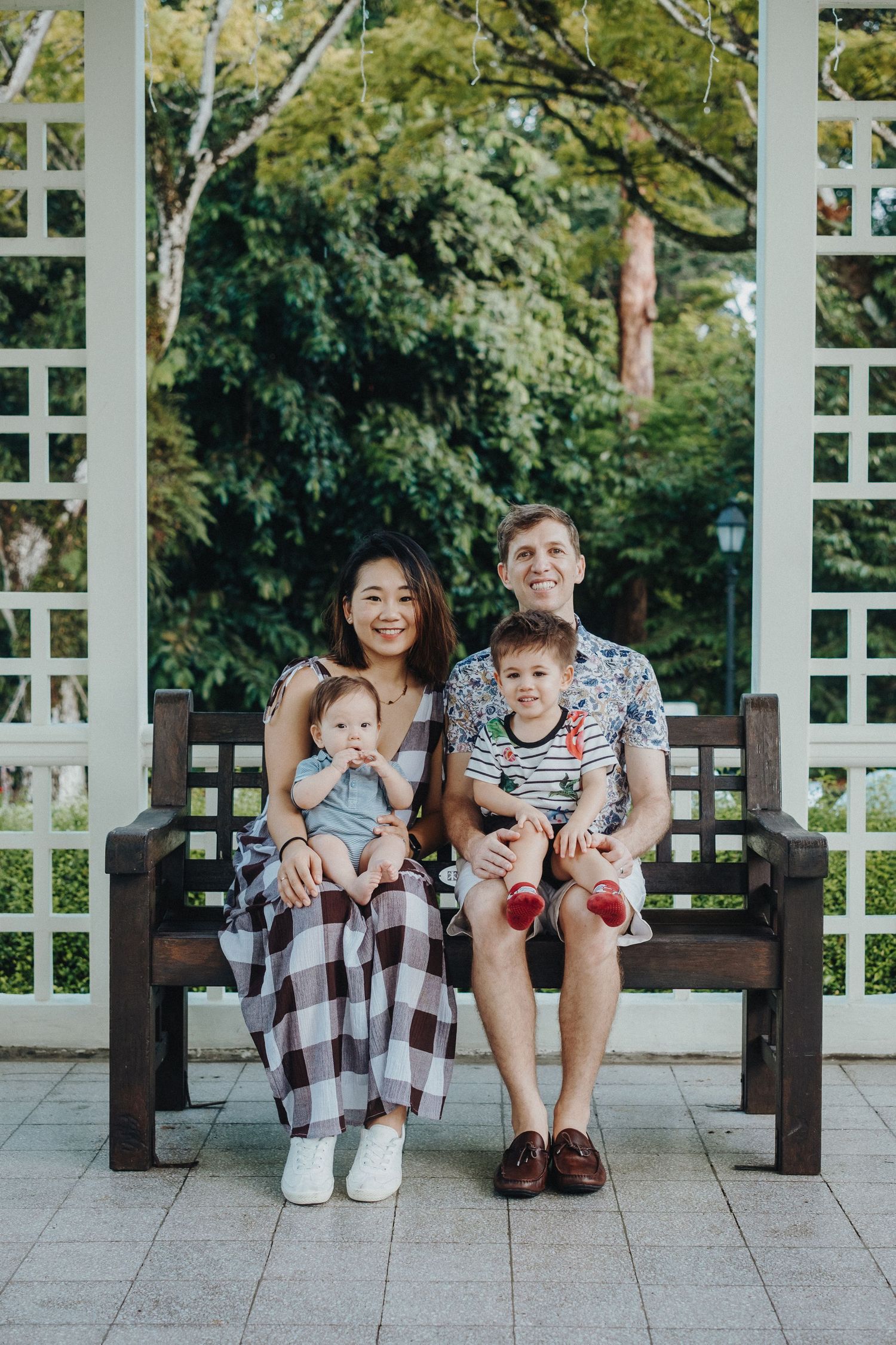 Family relaxing together on a wooden bench in front of a white garden trellis on a sunny day.
