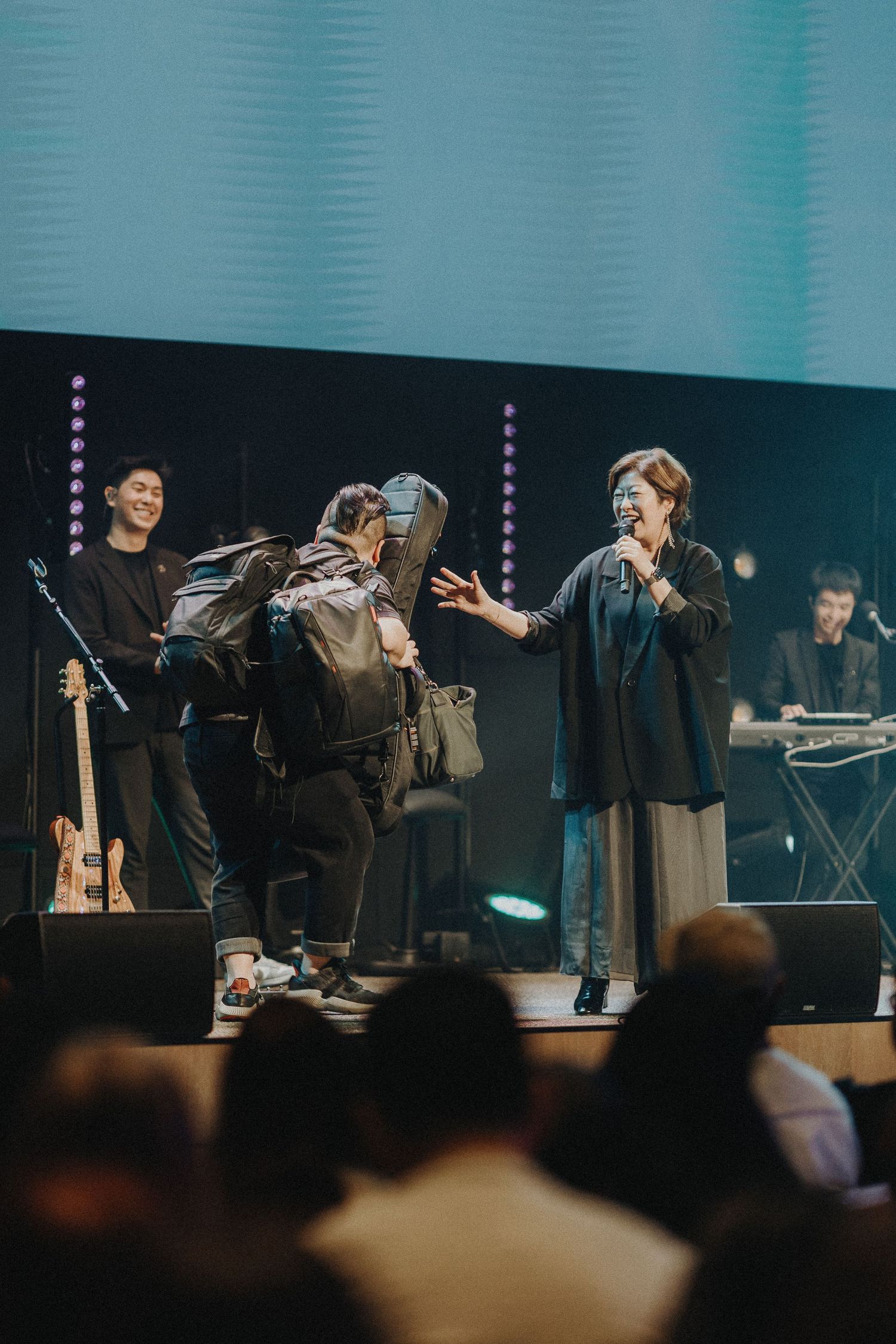 Musicians perform on a stage with turquoise lighting while band members interact during the live show.