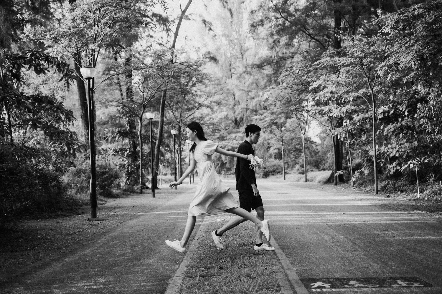 Two people playfully jumping and posing on a tree-lined path in black and white.