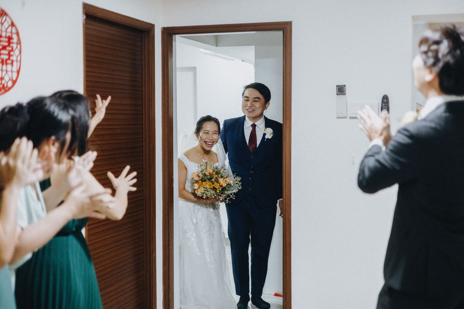 Wedding guests celebrate and wave as a newlywed couple enters through a doorway holding a yellow bouquet.