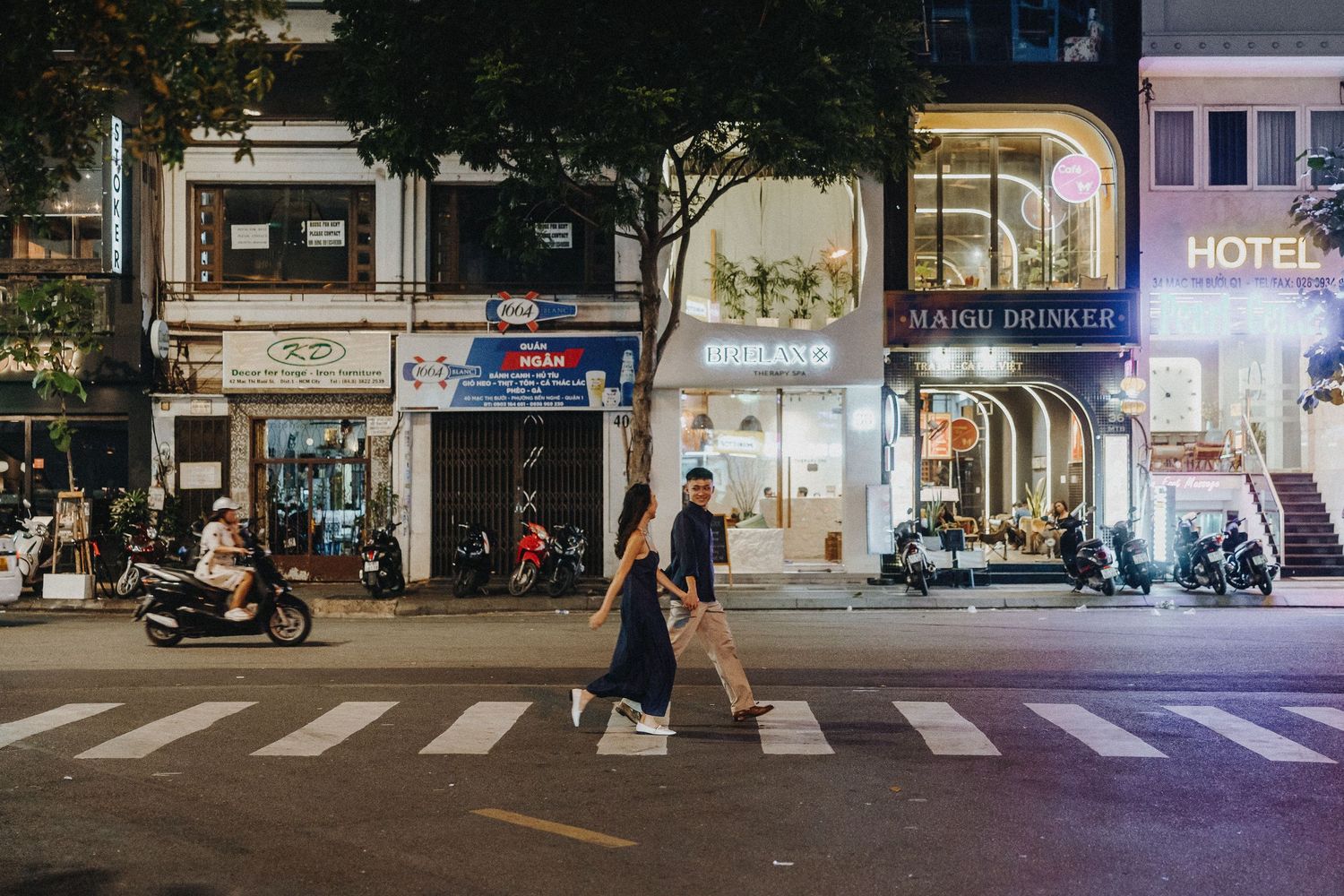 Person crossing a vibrant city street at night with illuminated storefronts and parked motorcycles in the background.