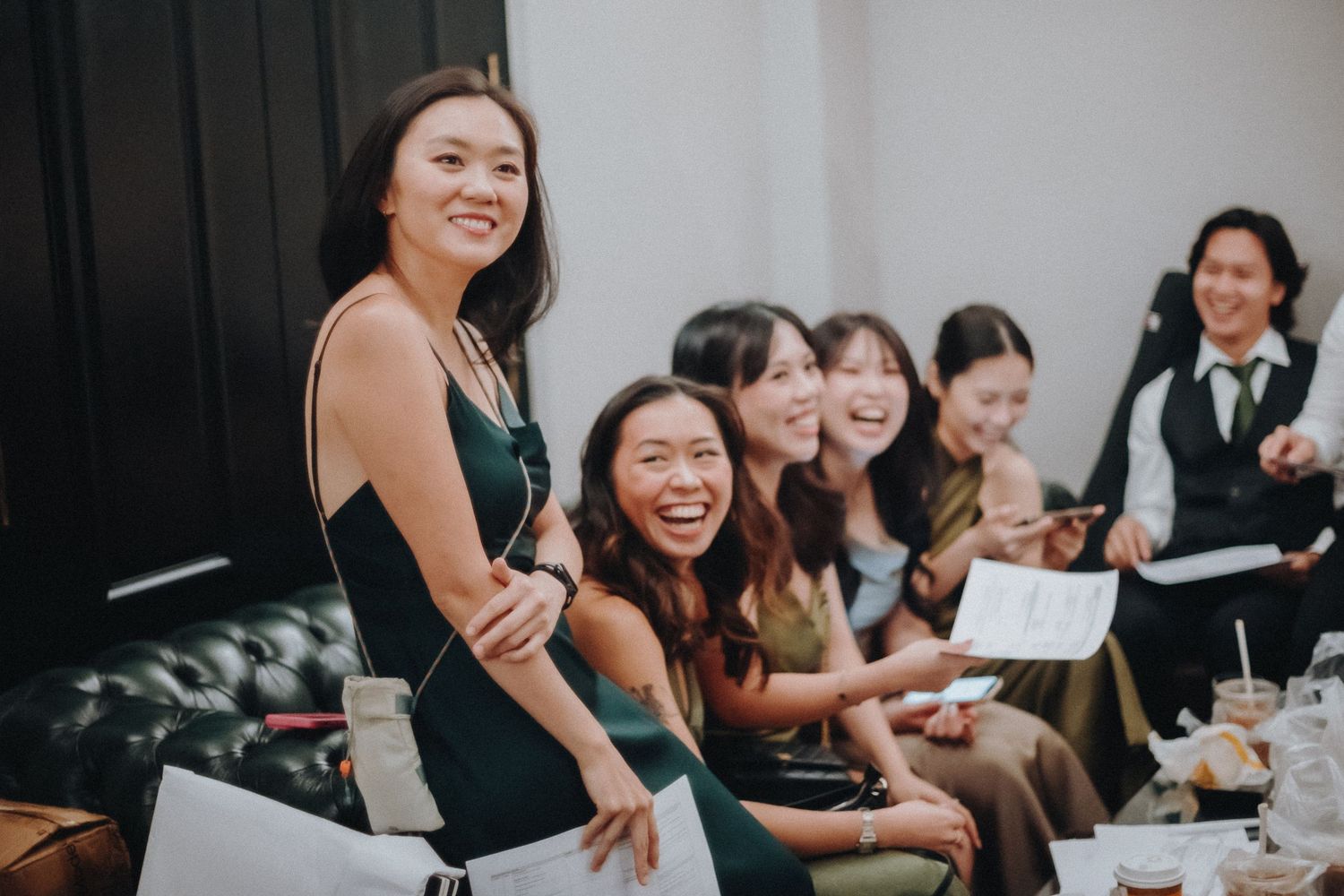 A group of friends in elegant attire share joyful laughter while gathered on a black leather couch at an indoor event.