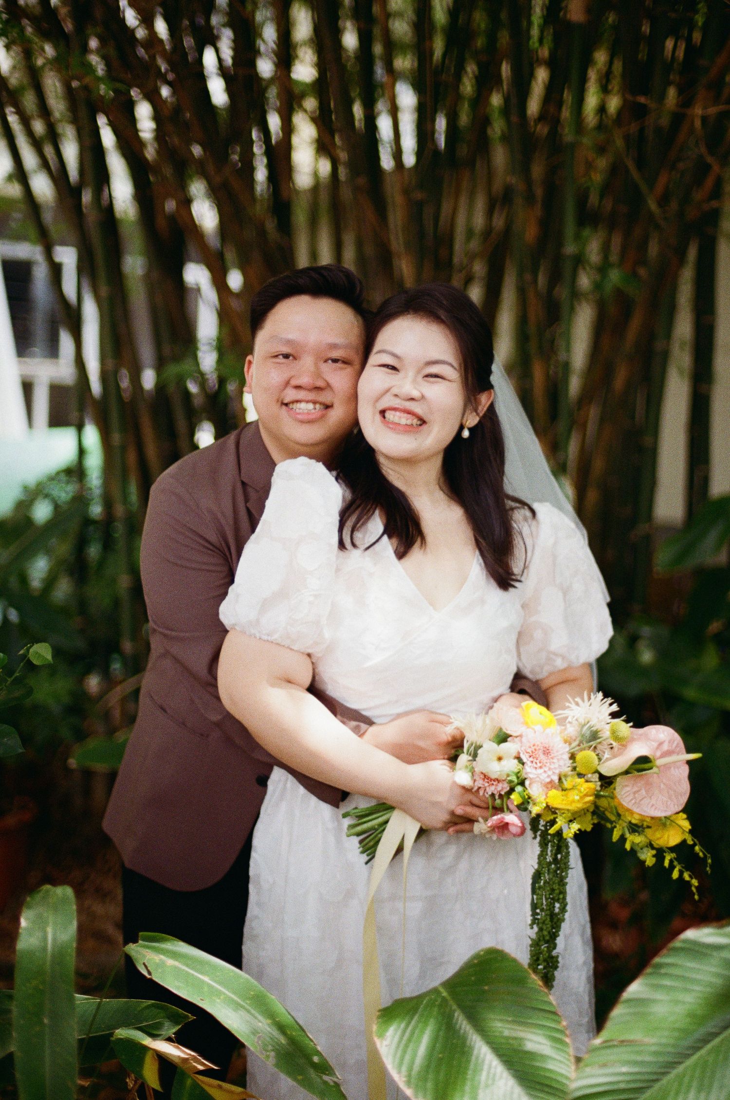 Two people pose affectionately outdoors in white clothing with a vibrant bouquet of flowers and tropical leaves.