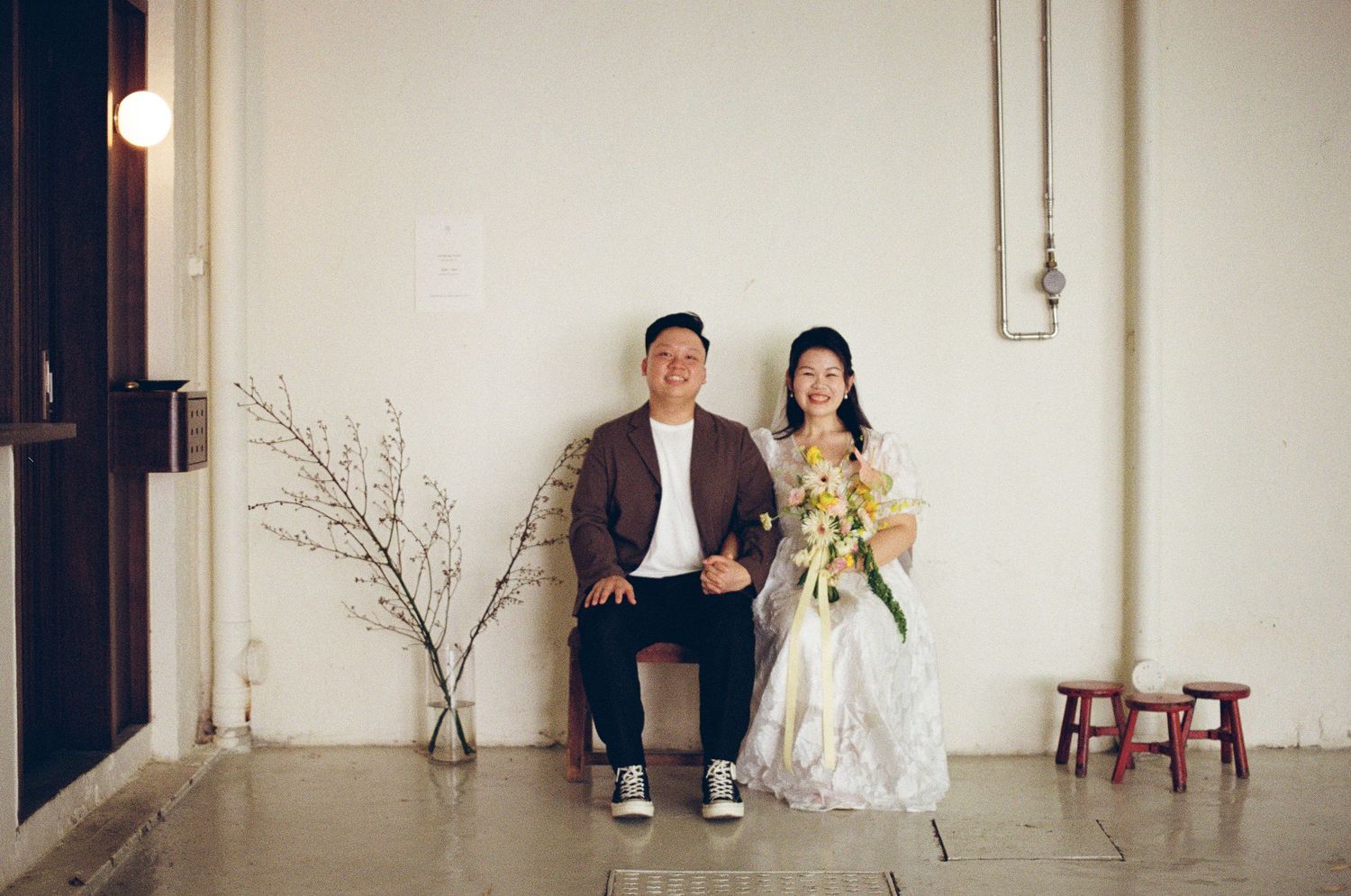 A couple in wedding attire poses for a romantic photoshoot against a white wall in a minimalist setting.