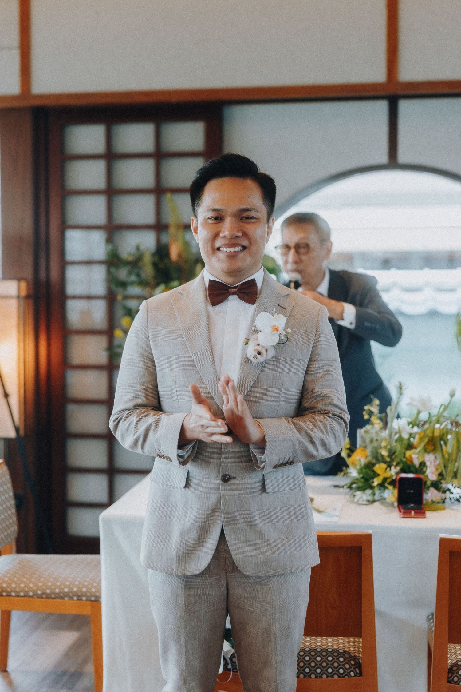A groom in a light gray suit and bowtie gestures during a wedding celebration in front of Japanese screens.