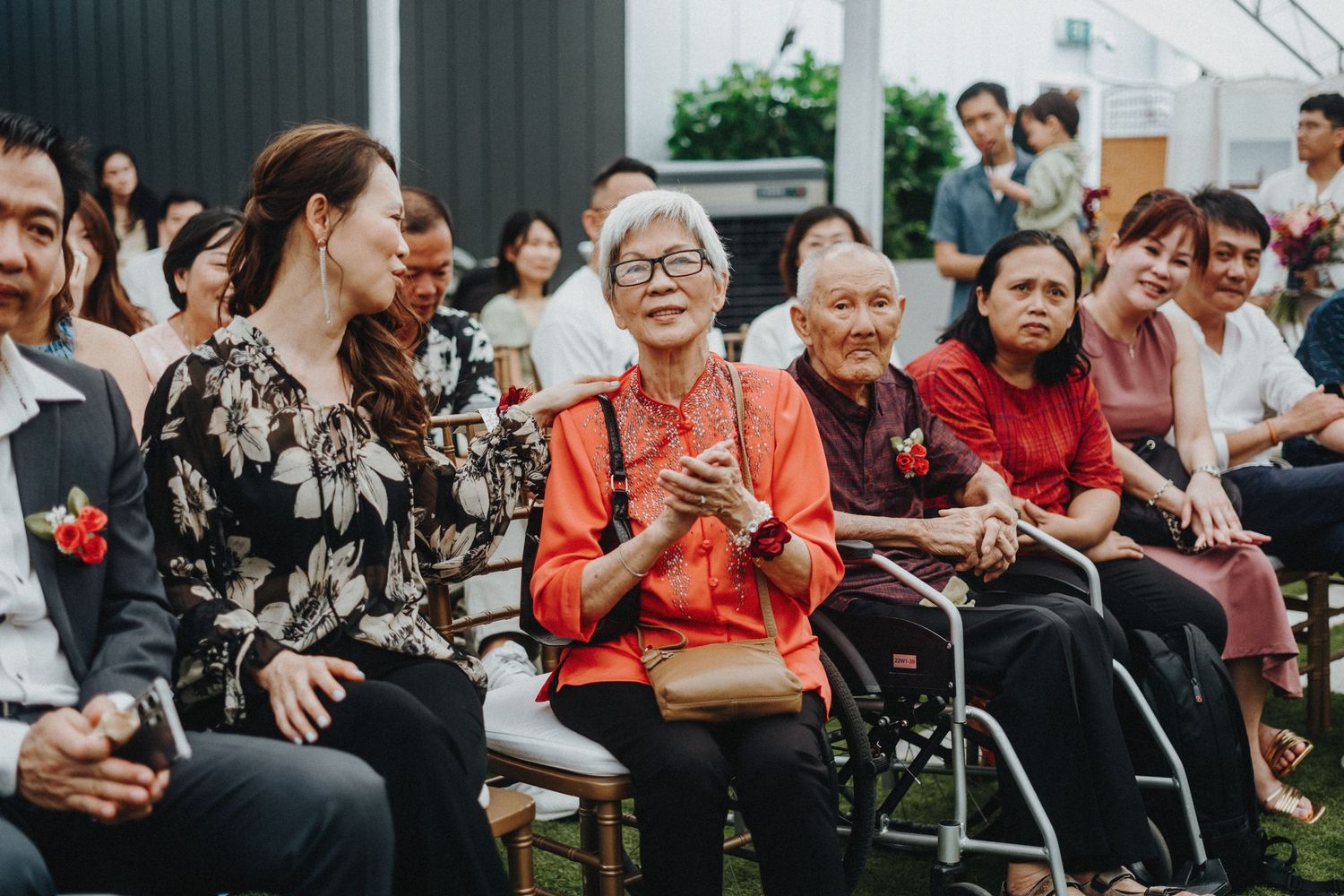 Wedding guests of various ages seated outdoors watching a ceremony with smiles and enthusiasm.