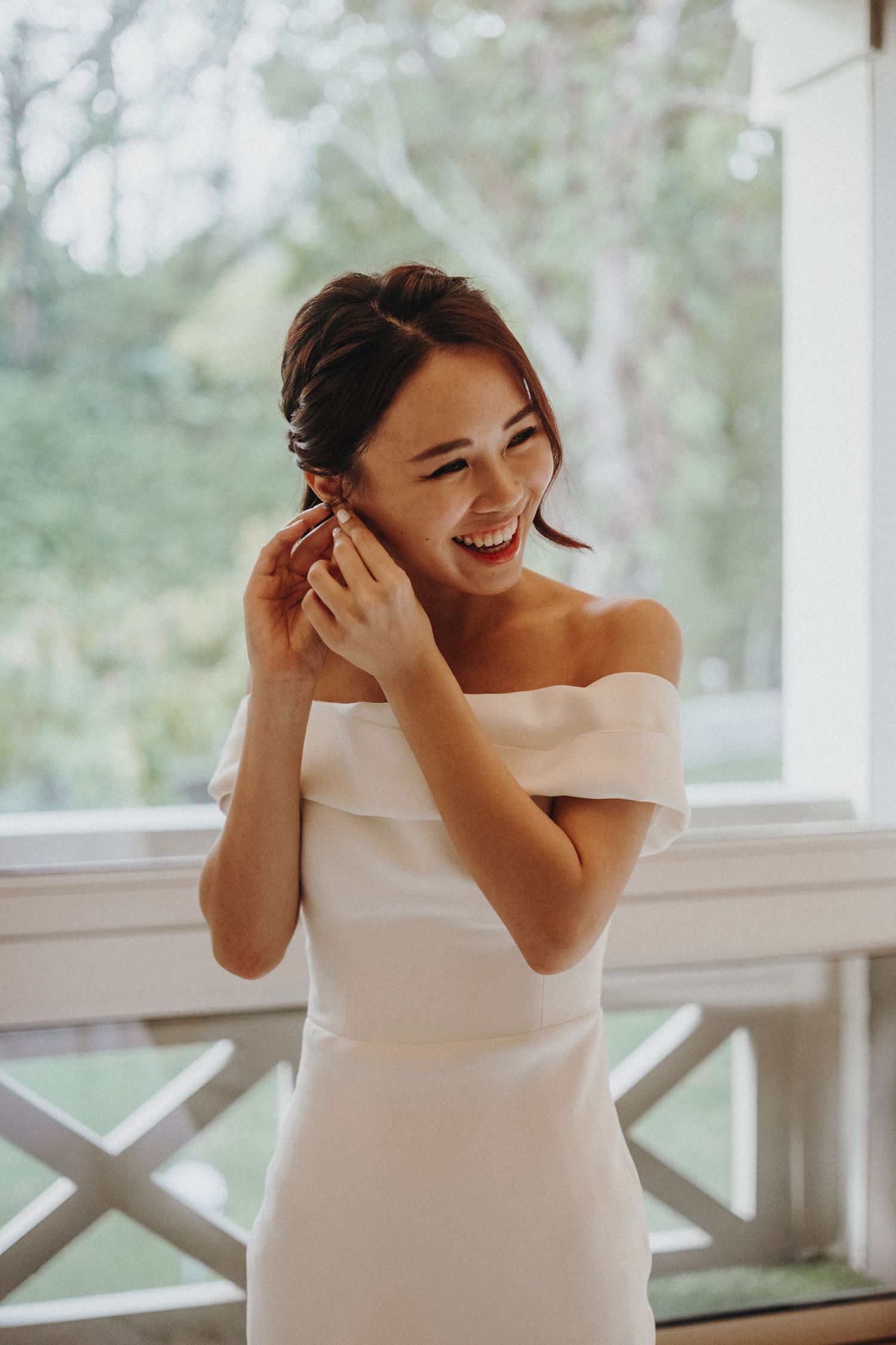 A person in a white off-shoulder dress joyfully covering their face while laughing.