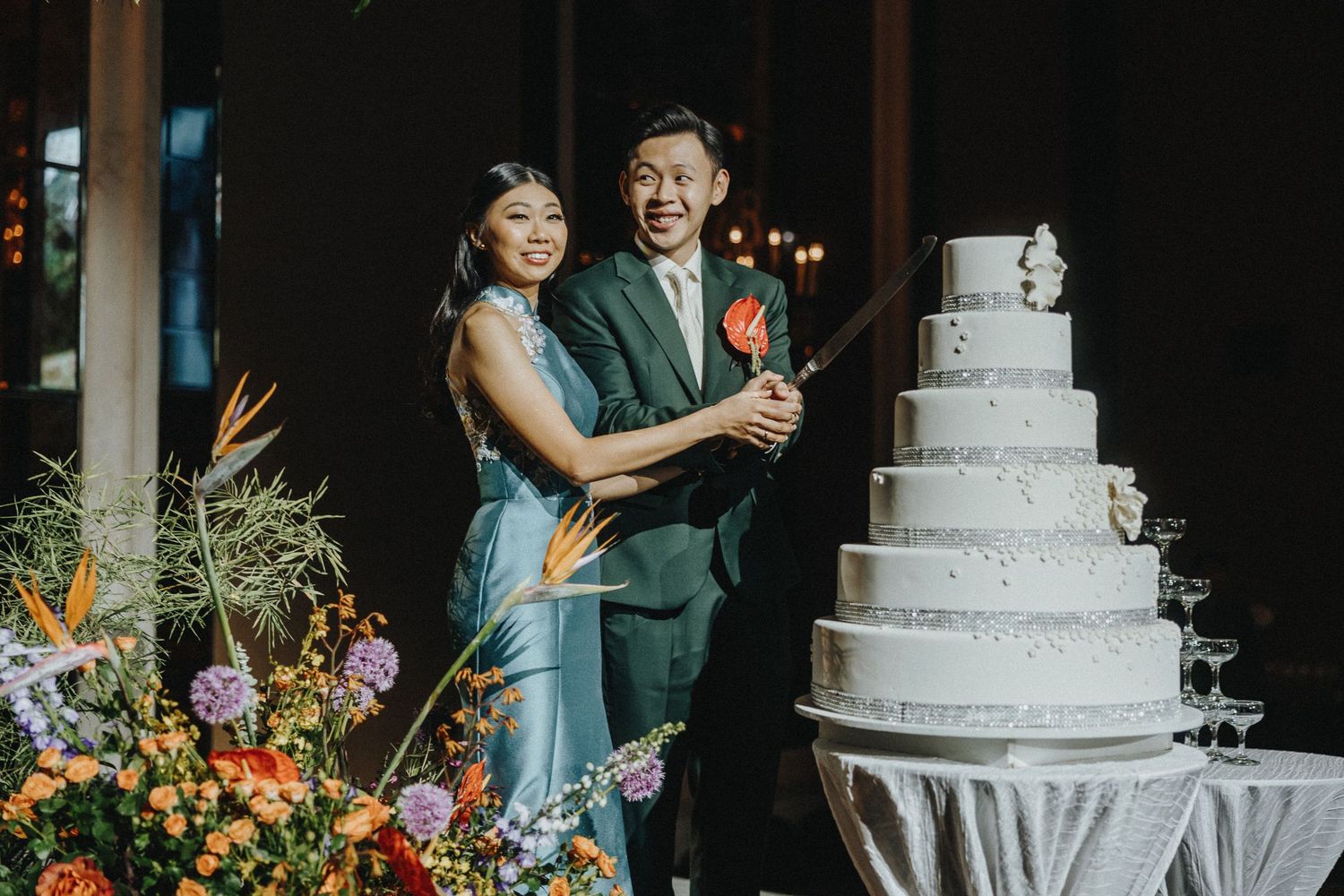 Wedding couple cutting elegant white tiered cake beside floral arrangements.