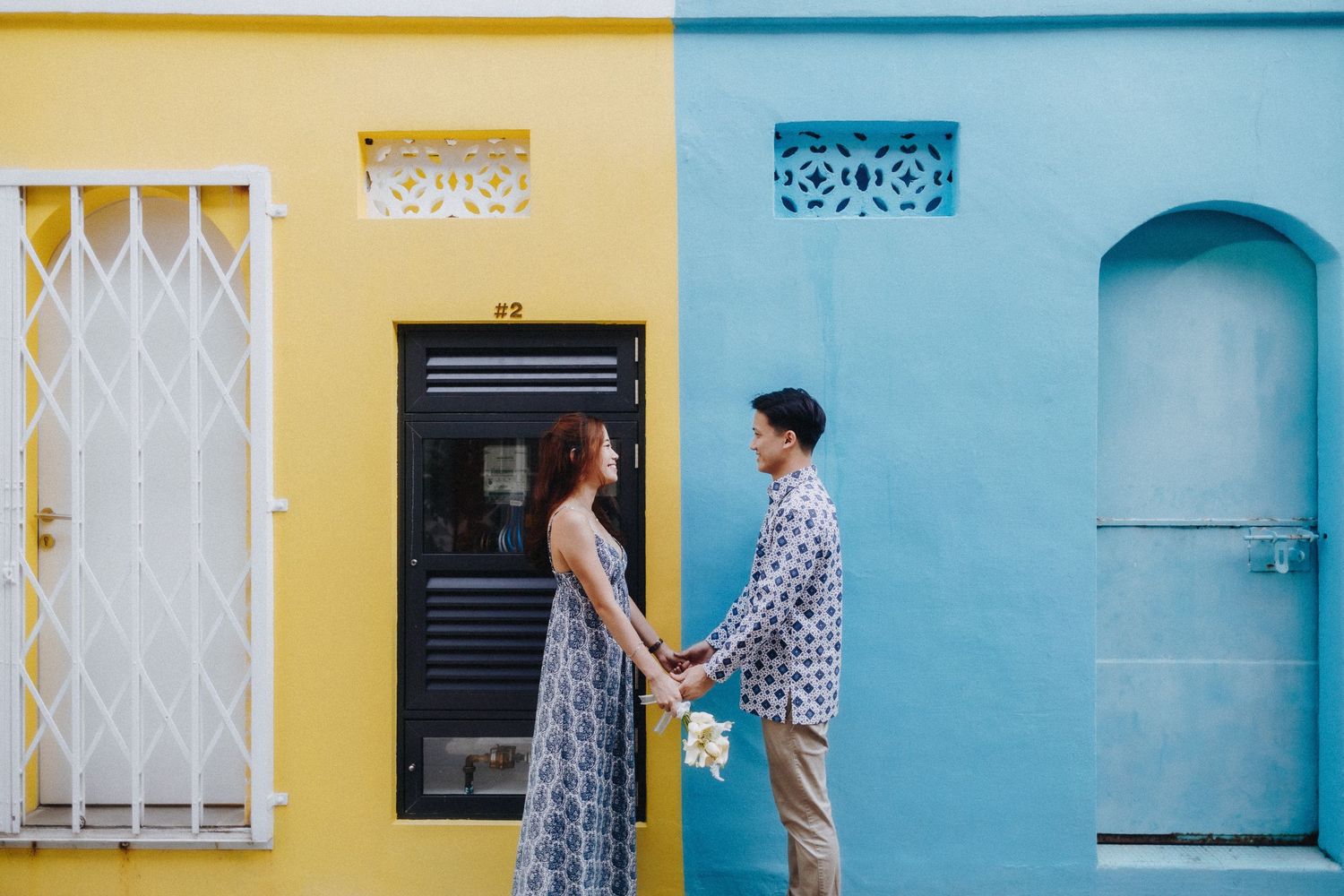 A couple stands in front of colorful yellow and blue building walls holding hands.