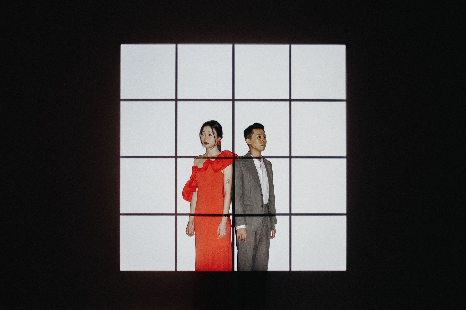 Two figures stand against a white gridded wall backdrop, one wearing red and one in gray formal attire.