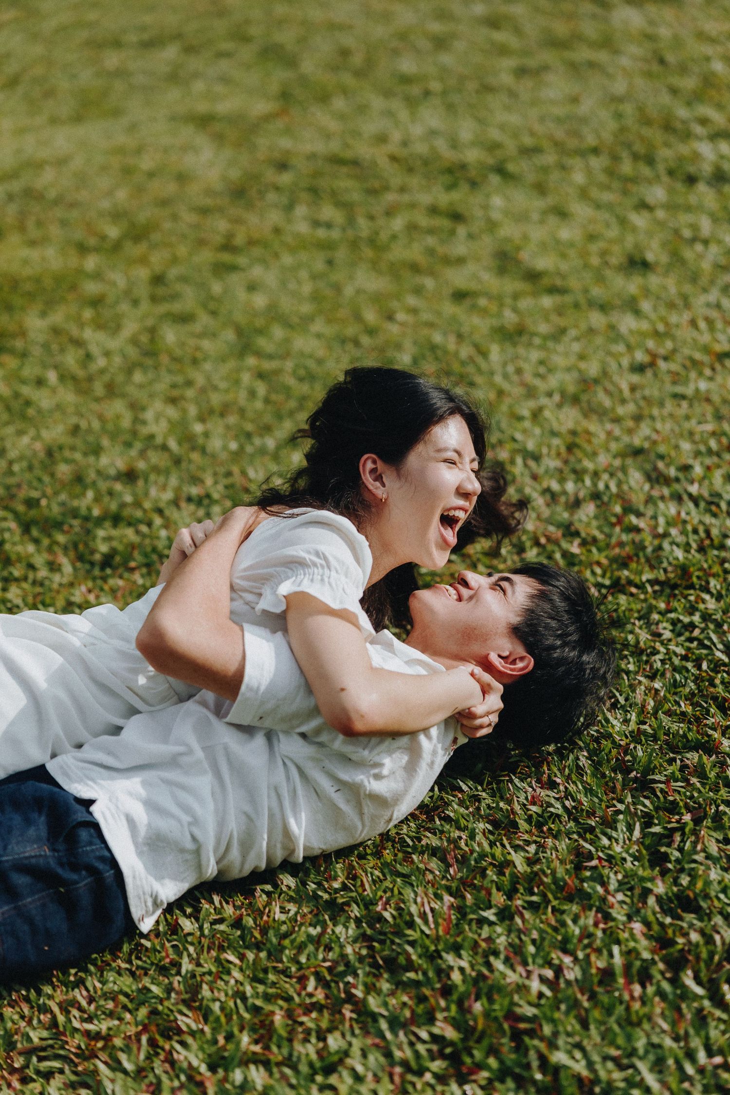 Playful couple laughing while lying on grass during outdoor photoshoot.