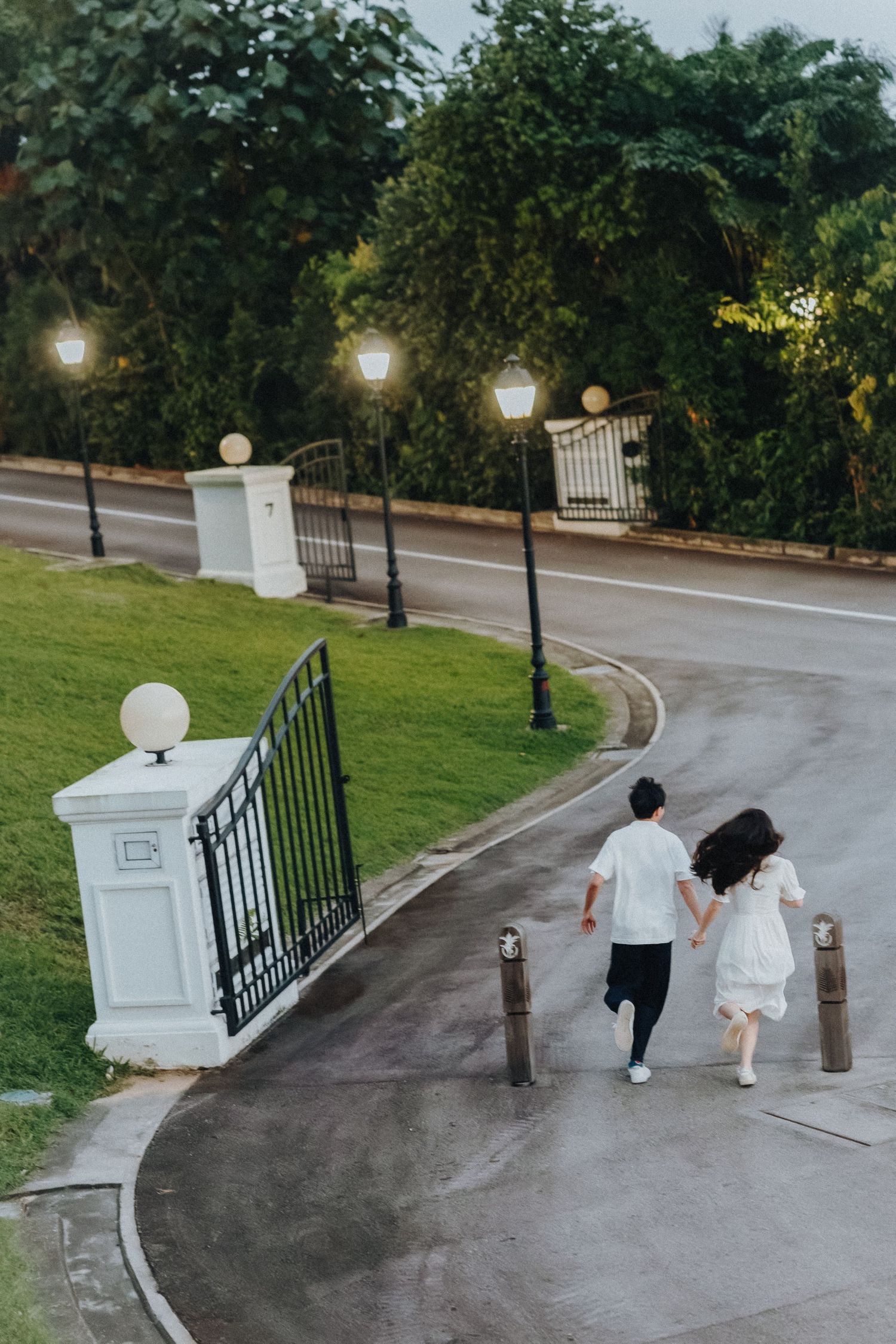 A couple walks down a curved garden path with vintage lampposts and iron gates at sunset.