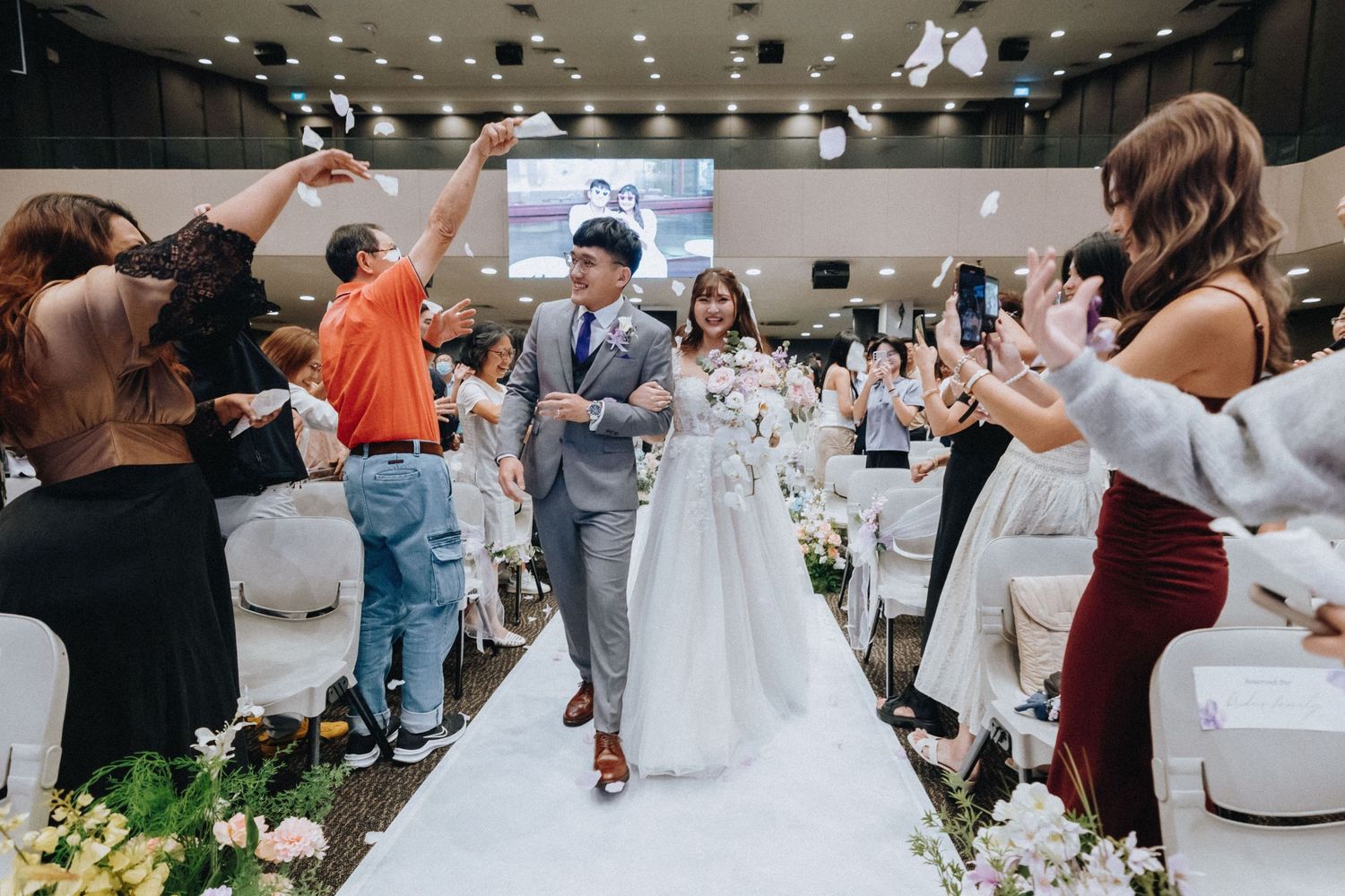 Wedding guests celebrate as the newlyweds walk down a white aisle runner at their reception.