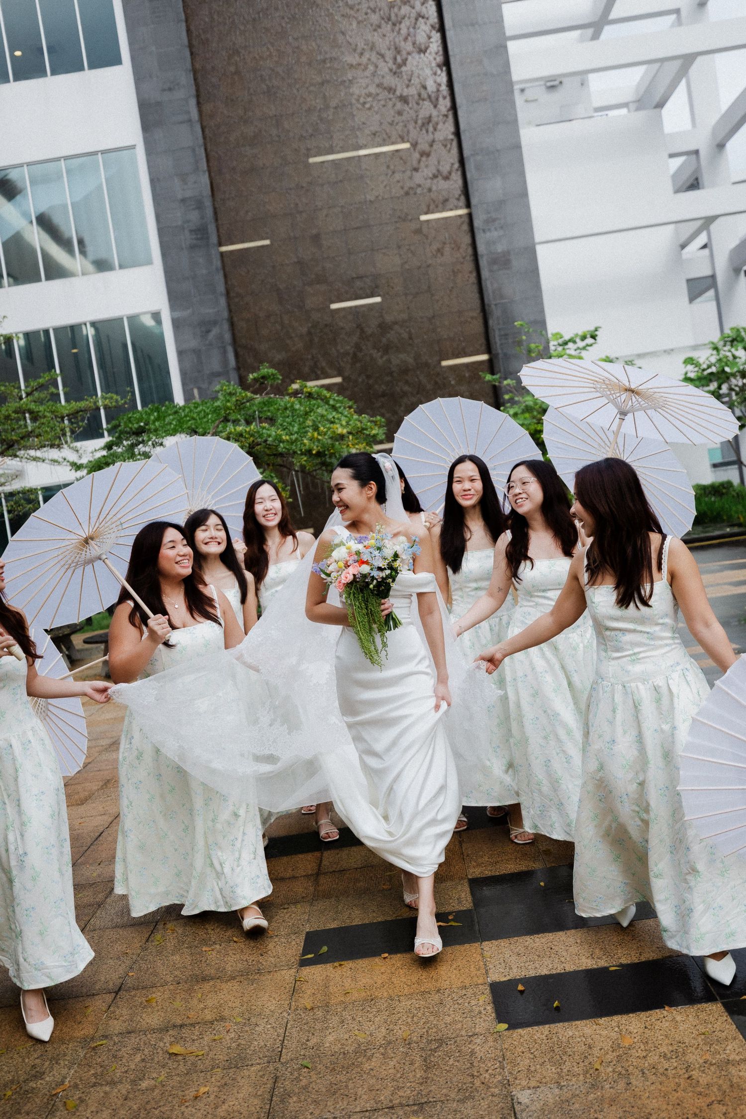 Joyful Asian bride walks with bridesmaids in matching white lace dresses carrying white parasols outdoors.