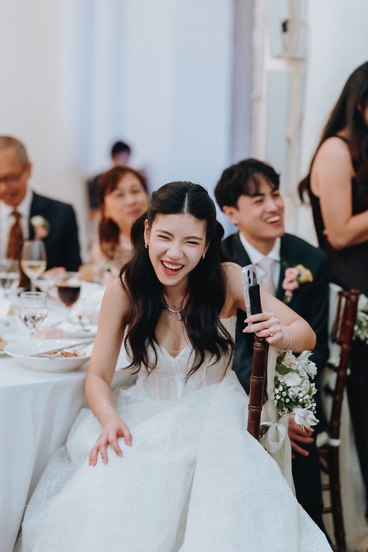Laughing bride in white wedding gown leans forward at reception table holding a microphone among smiling guests.