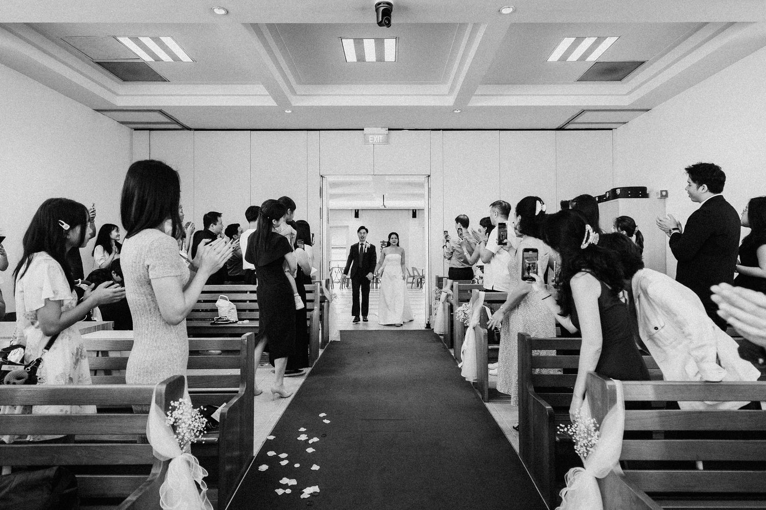 Black and white photo of a wedding ceremony with guests lining the aisle as the couple walks forward.