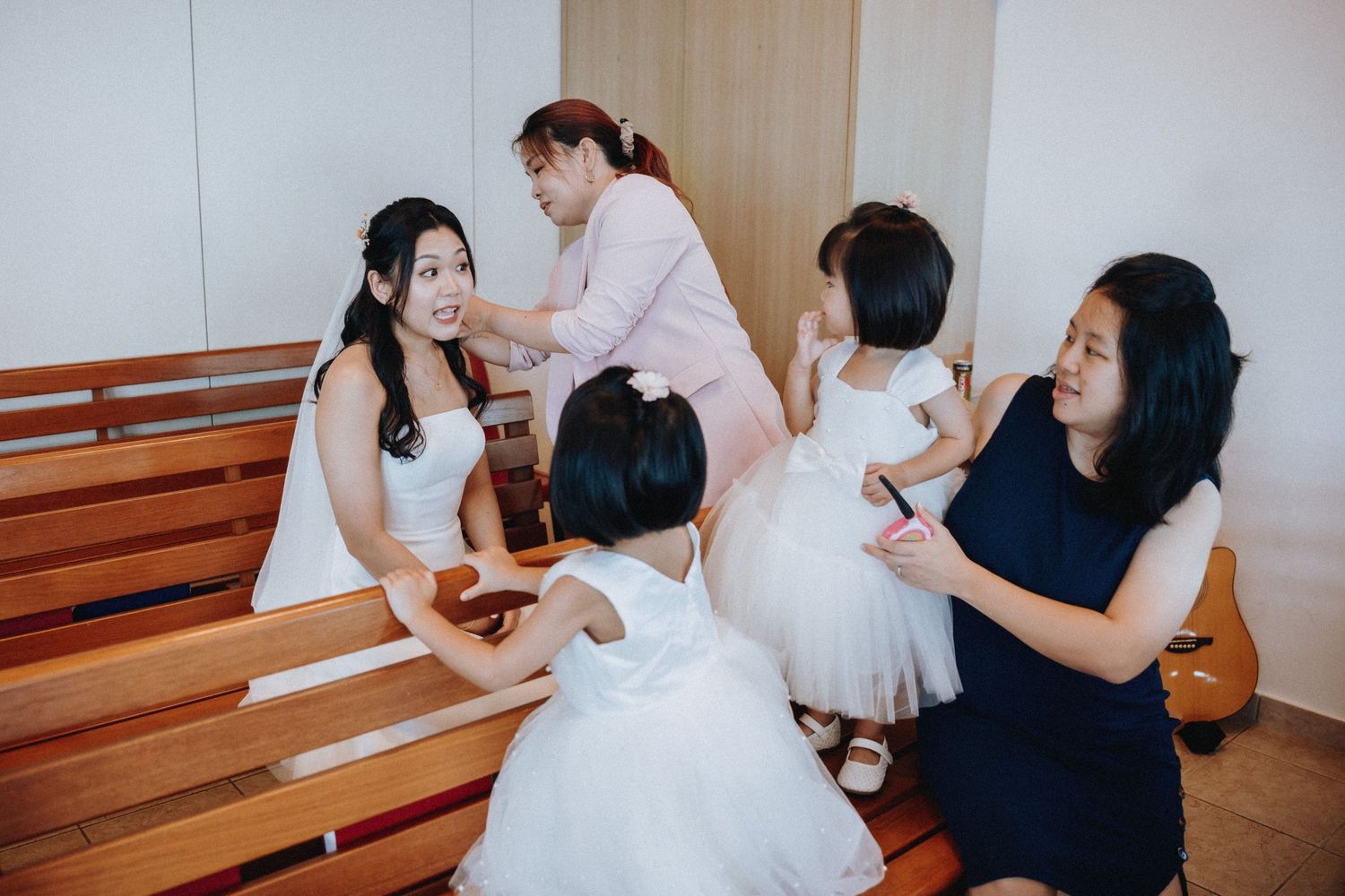 Bride in white dress sits with bridesmaids and flower girls during wedding preparations.