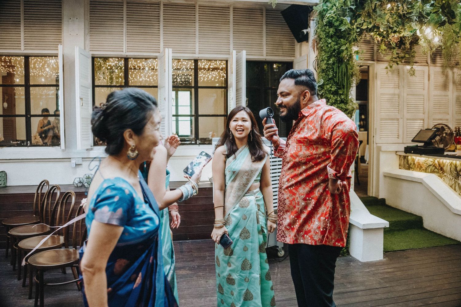 Guests laugh and chat with a man holding a microphone at an elegant indoor-outdoor wedding reception.