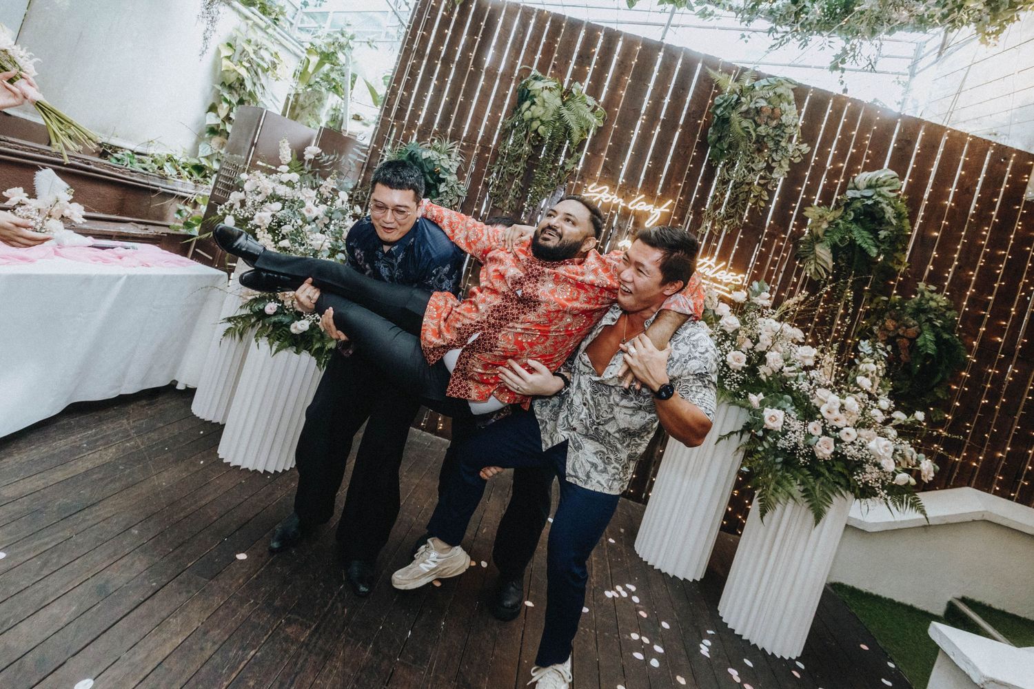 Three men playfully pose lifting one guest at a festive outdoor wedding reception.