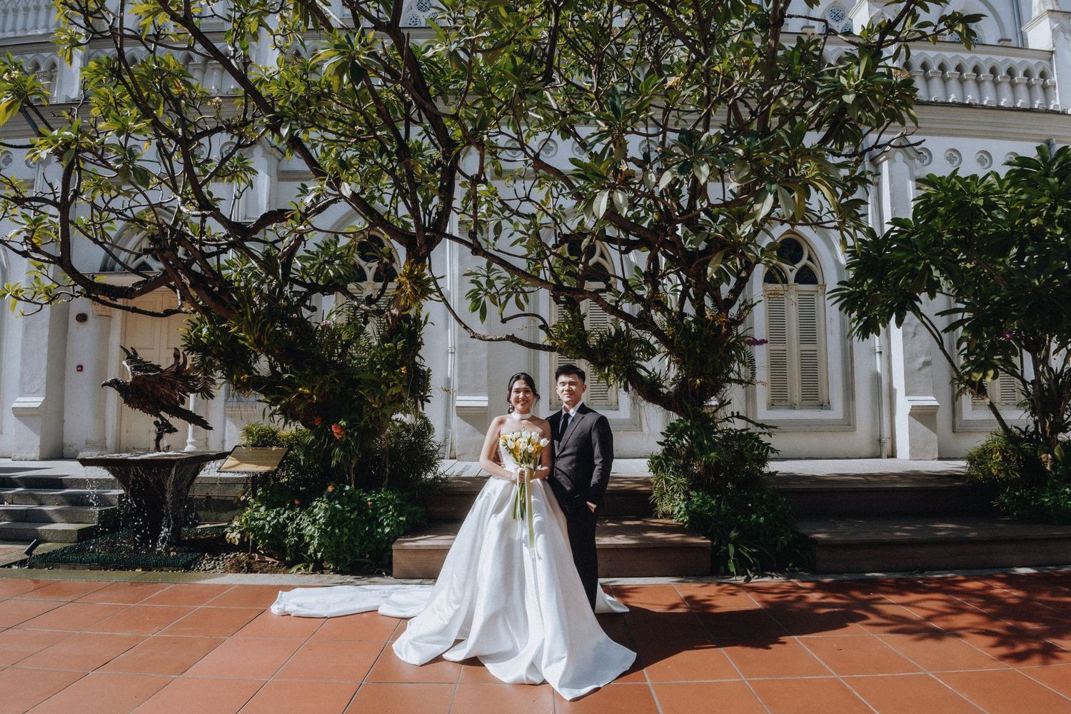 A bride and groom pose together in front of a historic white church surrounded by lush trees on their wedding day.