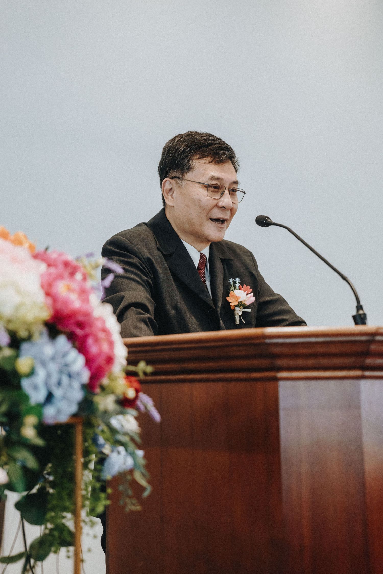 Older Asian man in dark suit speaking at wooden podium during wedding ceremony.
